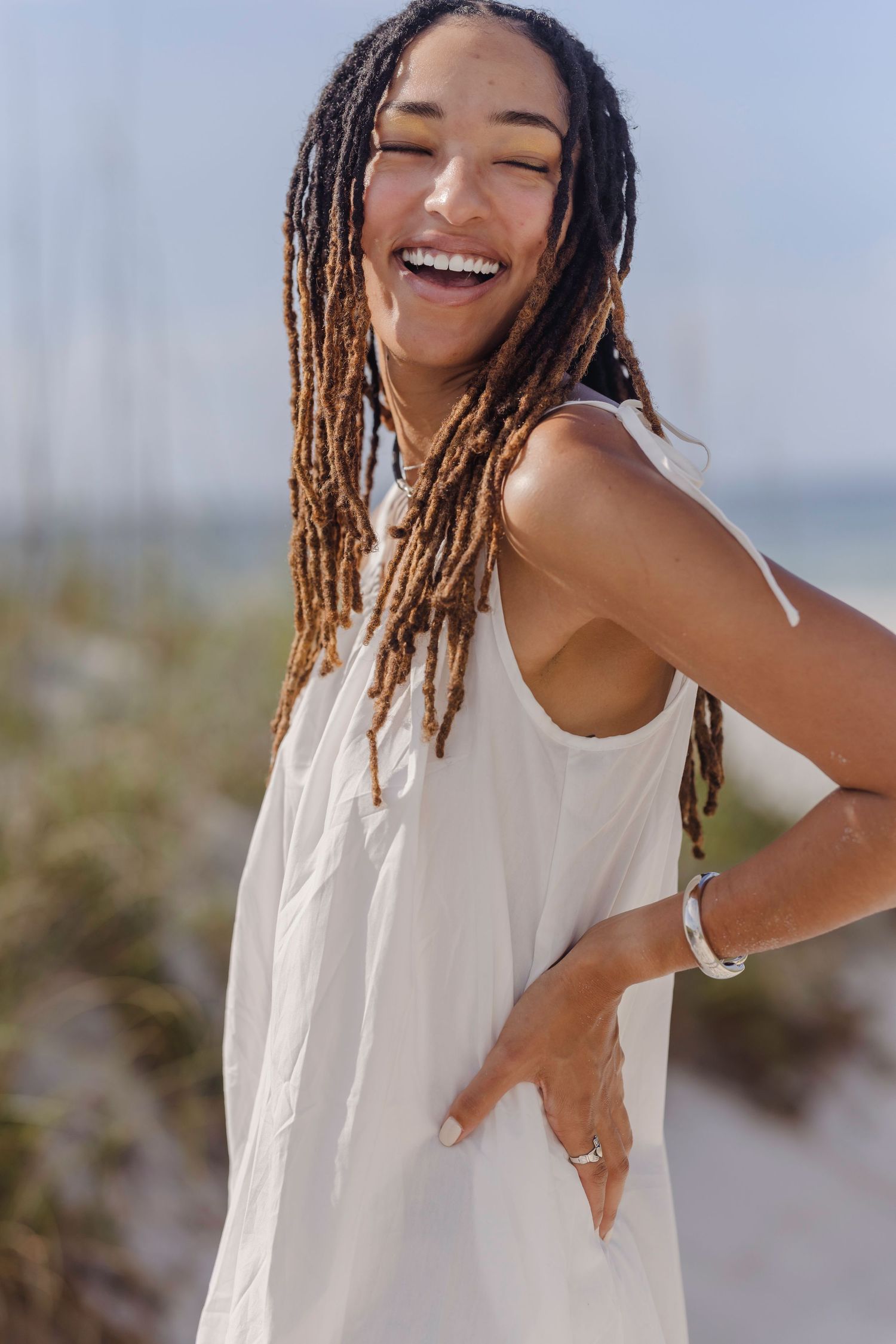 Smiling woman with long dreadlocks wearing a white sundress at the beach on a sunny day.