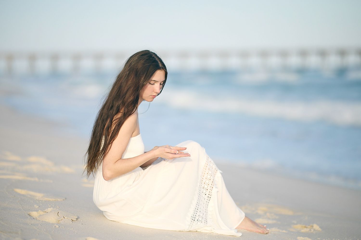 A young woman in a white dress sits peacefully on a sandy beach, gazing down with long dark hair flowing.