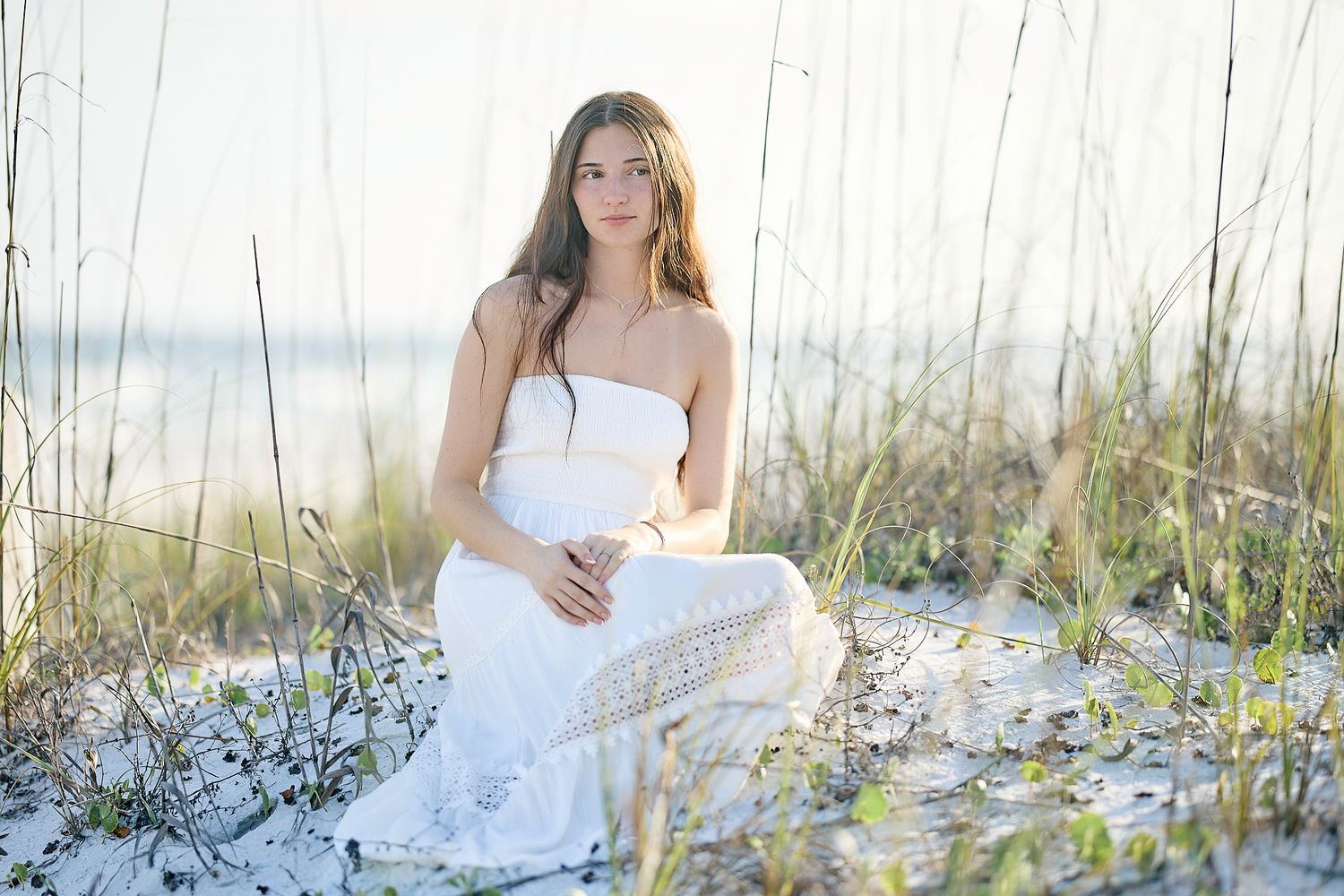 Young woman in white dress sitting on sandy beach dunes with sea oats, serene coastal portrait photography.