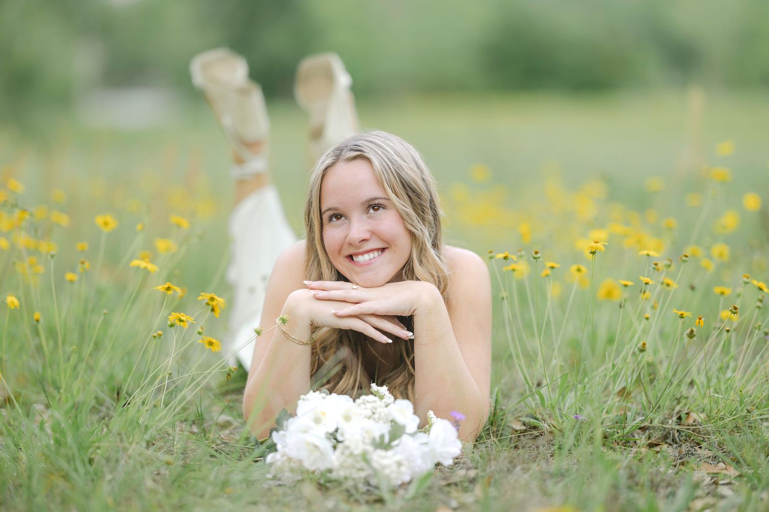 Smiling blonde woman lying in a wildflower field holding white flowers, surrounded by yellow blooms in a lush green meadow.