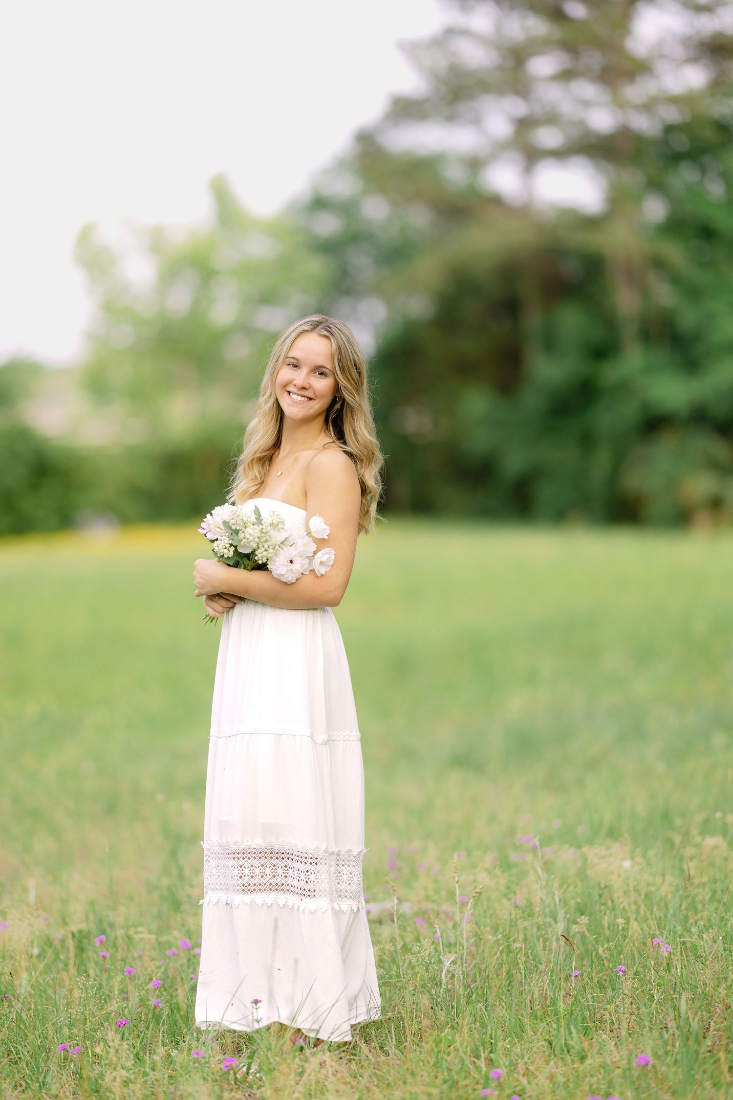 Young blonde woman in white strapless maxi dress holding pink flowers in a lush green field.