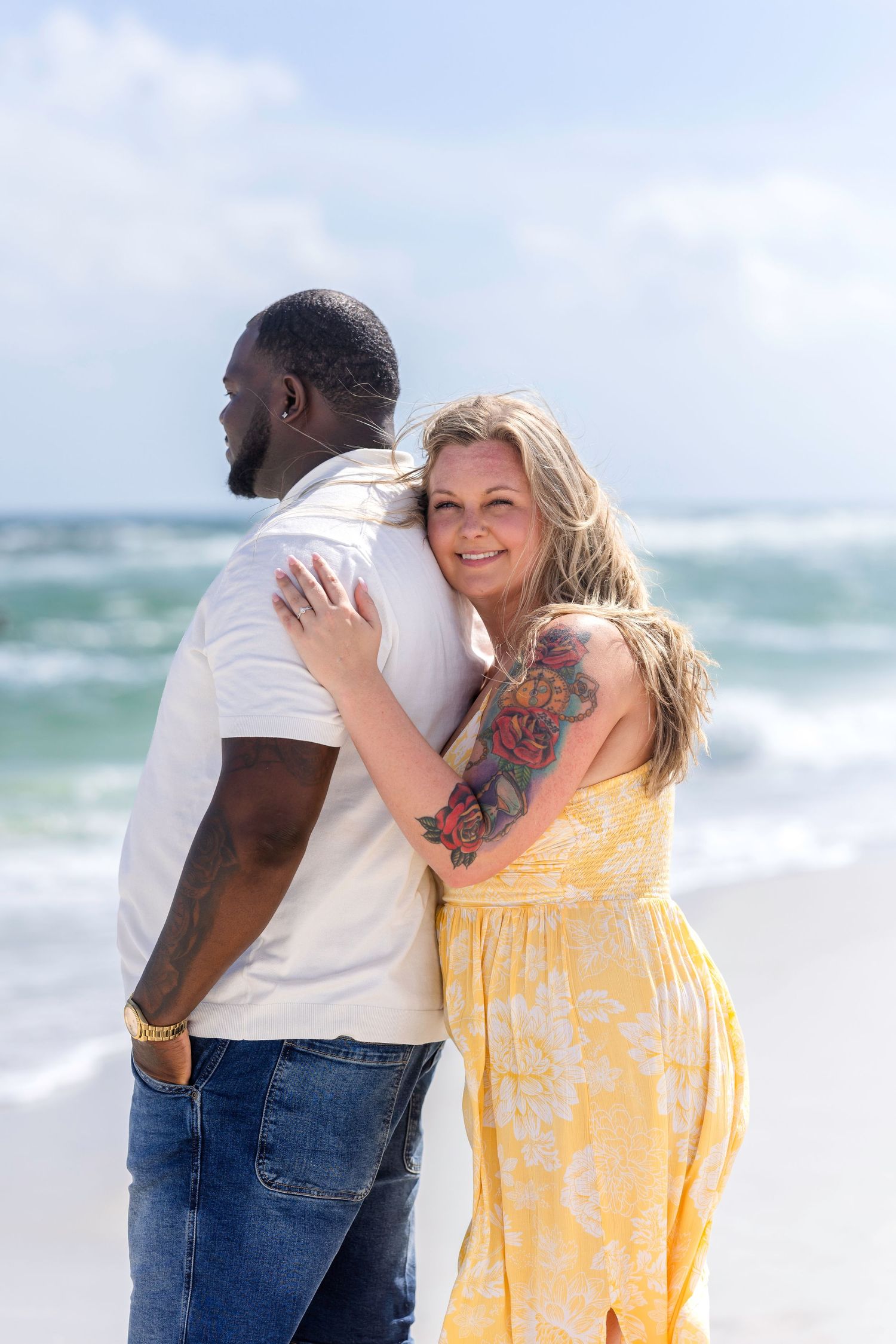 Woman in yellow floral dress with tattoos embraces man from behind on a sunny beach by the ocean.