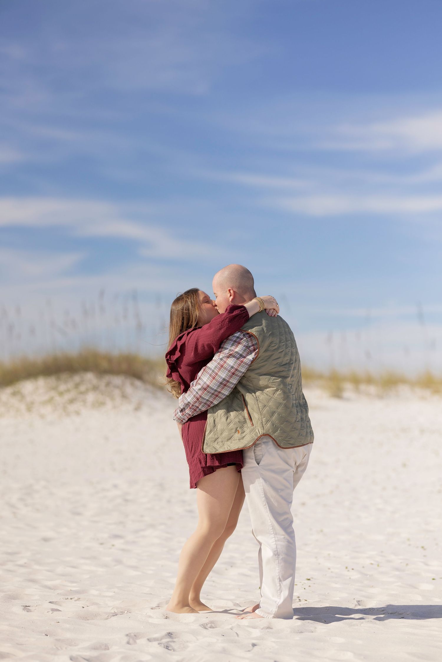Couple embracing on white sand beach dunes under blue sky, woman in burgundy dress and man in plaid shirt and green vest.