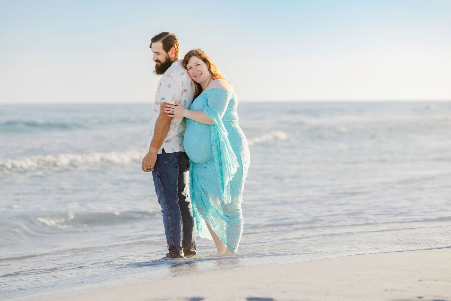 Pregnant couple embracing on a beach, woman in teal dress, waves in background, maternity photo session.