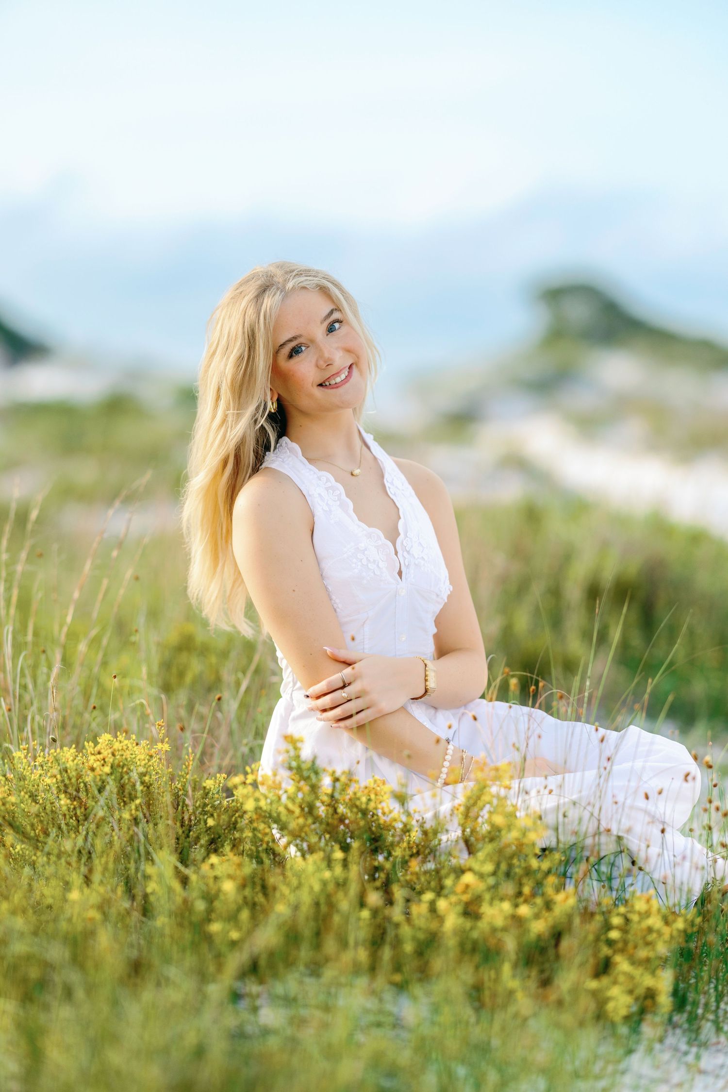 Smiling blonde woman in white dress sitting in green meadow with yellow wildflowers on a sunny day.