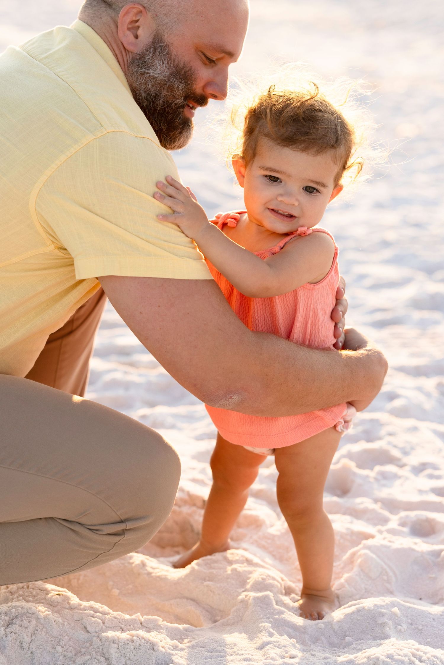 A bearded father in a yellow shirt helps his toddler daughter stand on a sandy beach during golden hour.