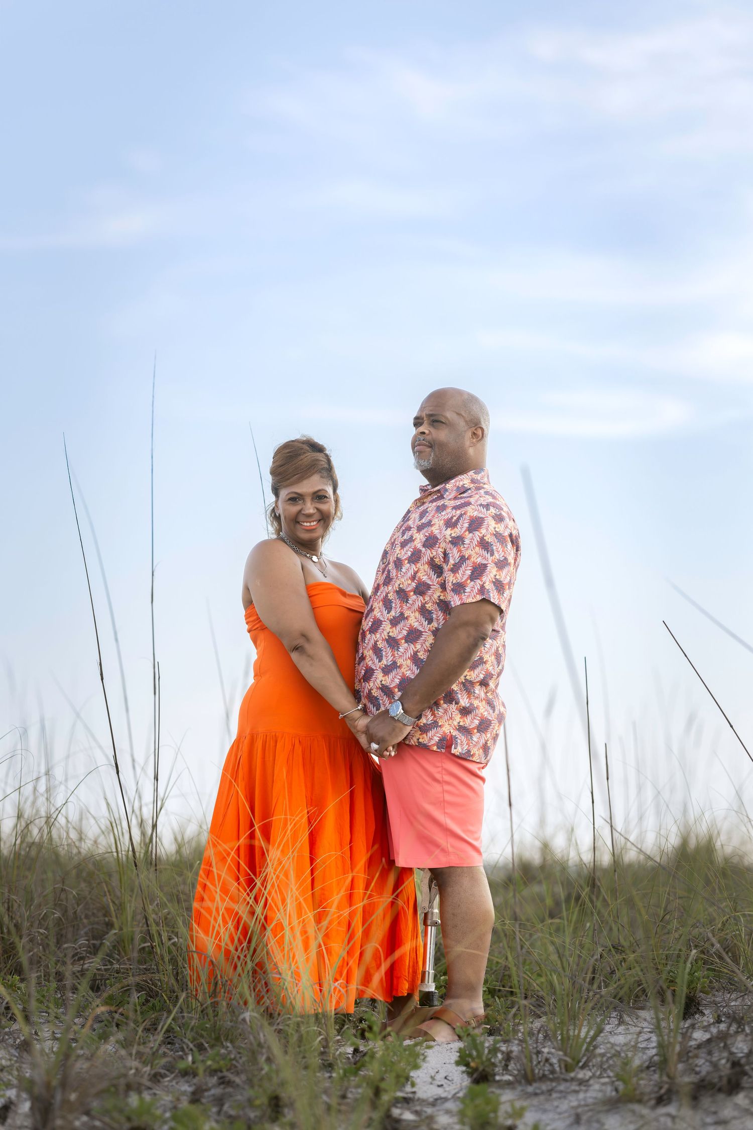 A couple holds hands and smiles among beach grass, woman in orange dress, man in pink floral shirt and coral shorts.