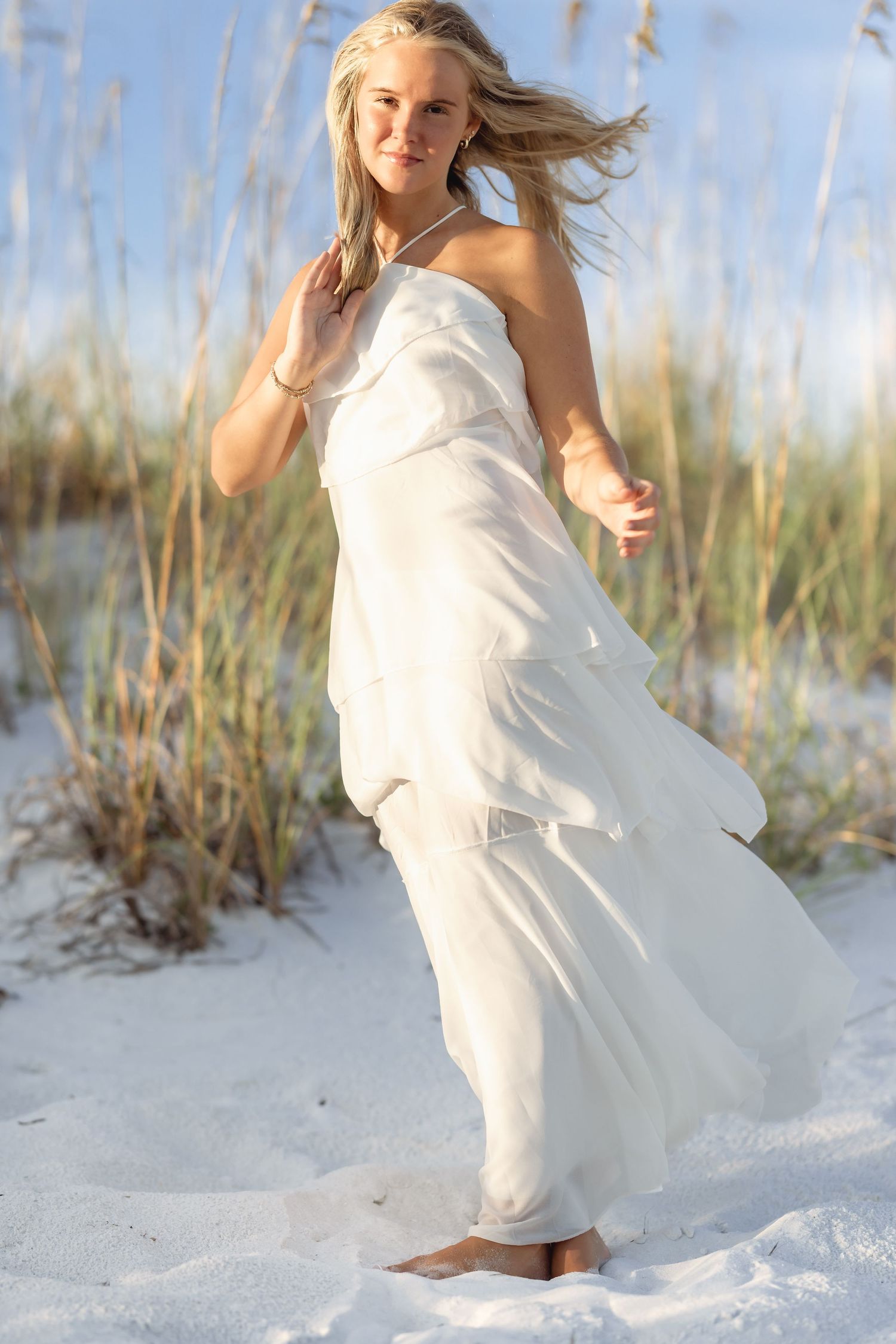Young blonde woman in flowing white dress standing barefoot on white sand beach dunes with sea oats in the background.