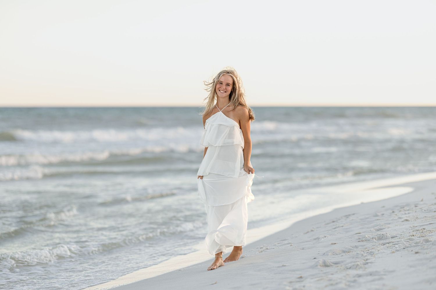 Blonde woman in white strapless maxi dress walking barefoot on sandy beach with ocean waves behind her.