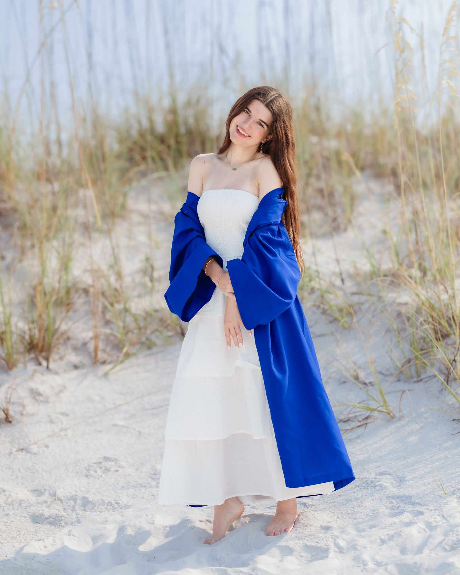 Young woman in white dress and royal blue coat posing barefoot on sandy beach dunes.