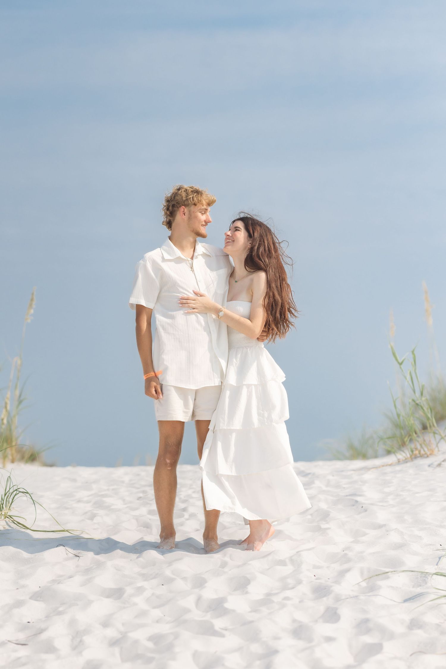 Young couple in white outfits sharing a romantic moment on a white sand beach with sea oats and blue sky.