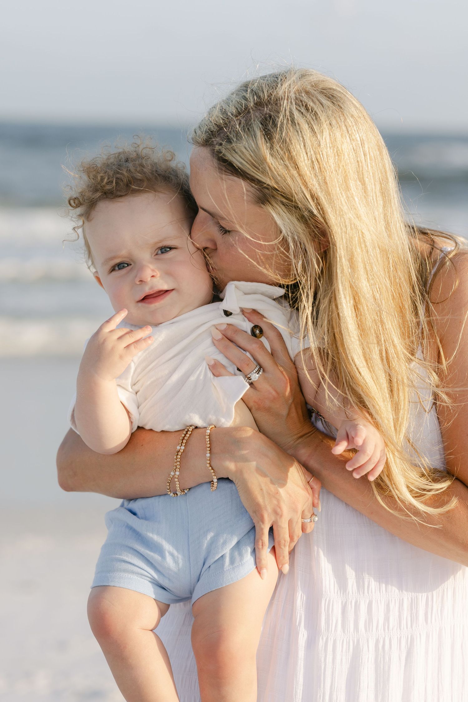 A mother tenderly kisses her curly-haired baby on a sunny beach, both dressed in white and blue.