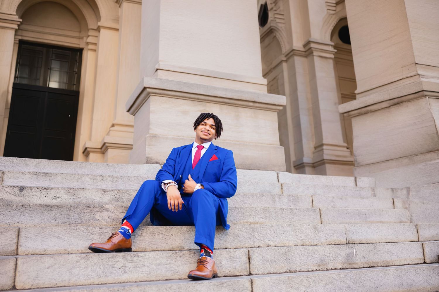 Man in blue suit with red tie sitting on marble steps of a grand building, wearing brown leather shoes.