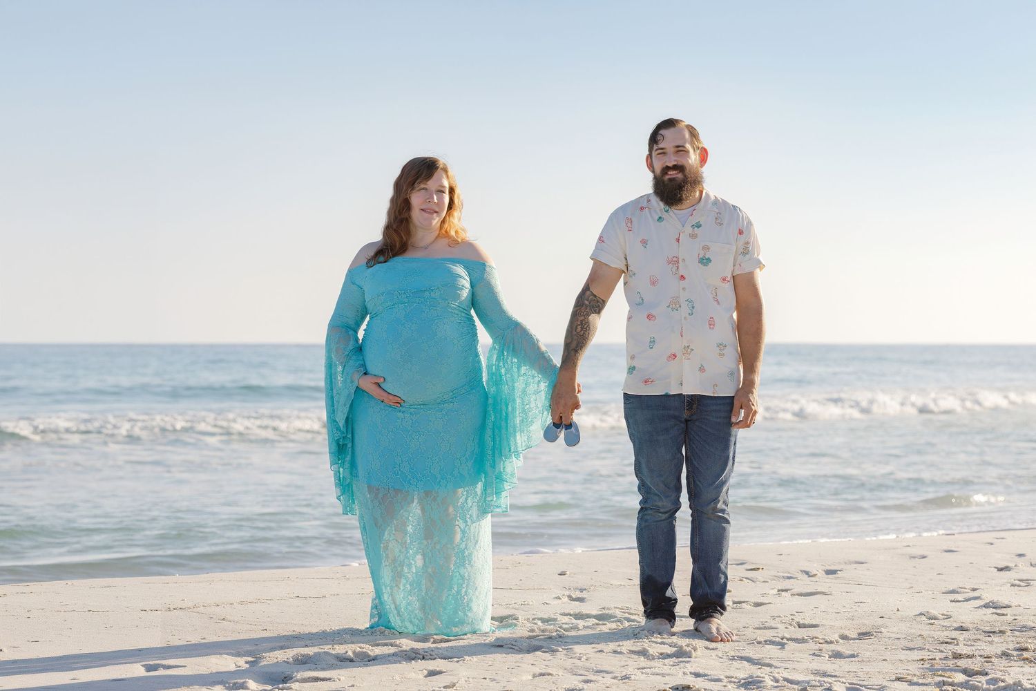 Expecting couple holding hands walking on beach, woman in flowing turquoise maternity dress.