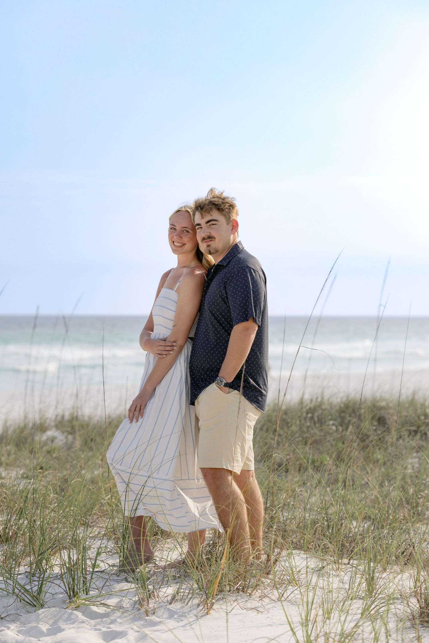 A couple embraces on a sandy beach dune with sea oats, ocean waves visible in the background on a sunny day.