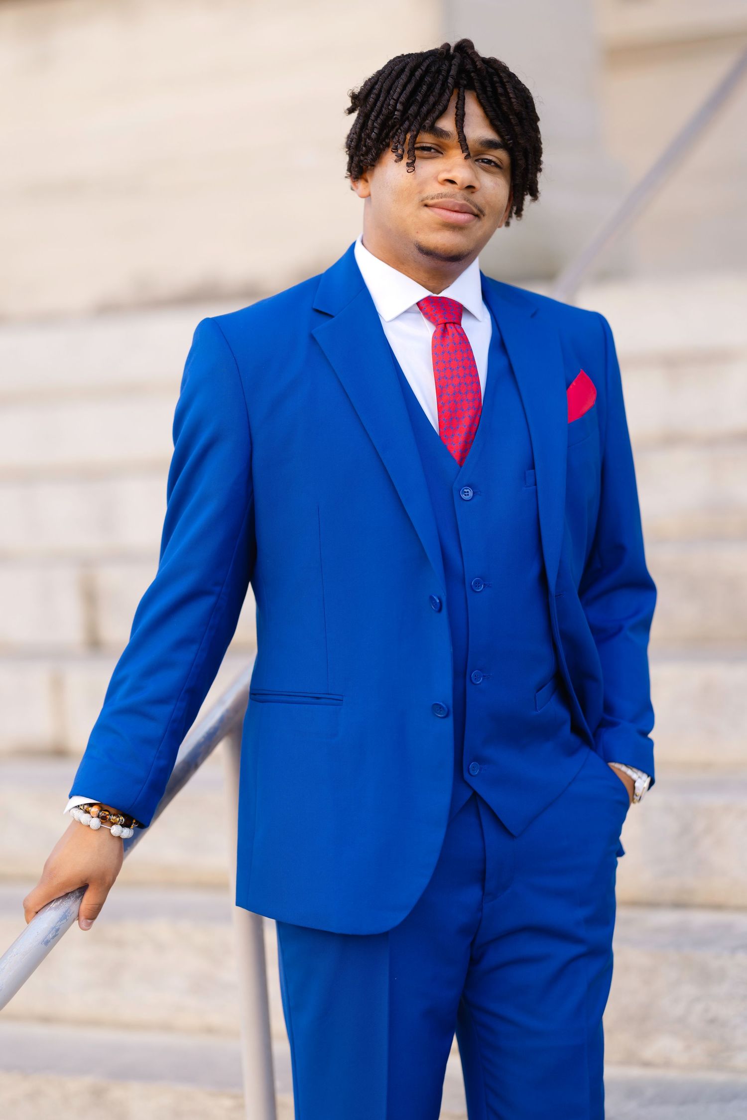 Young man in a royal blue three-piece suit with red tie and pocket square, posing on outdoor steps.