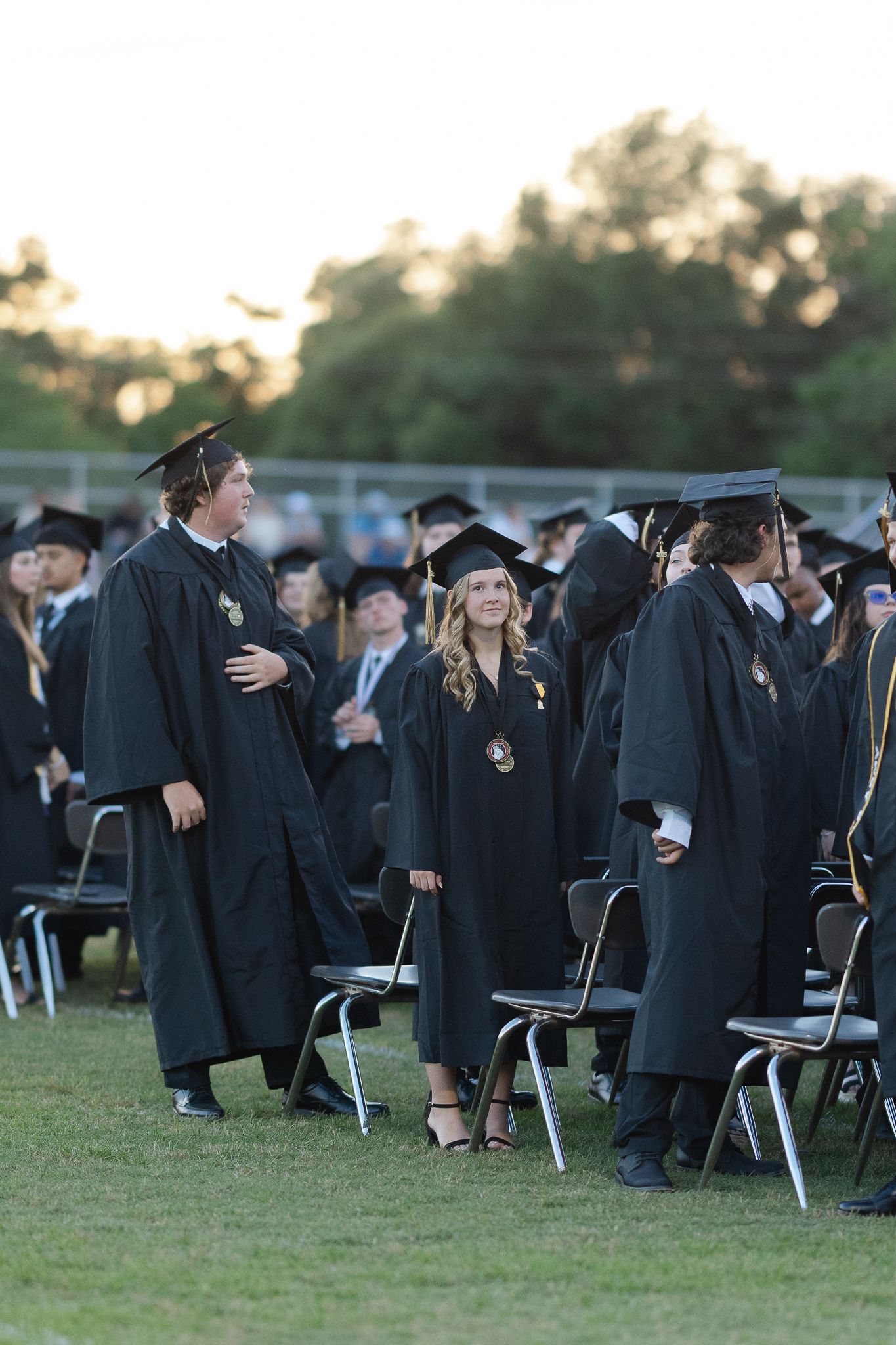 Graduates in black caps and gowns stand among rows of chairs during commencement.
