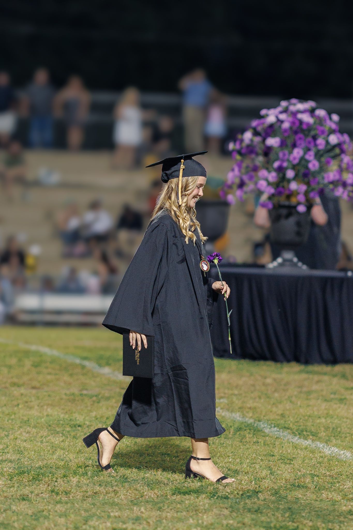Graduate walks past purple flowers in traditional black graduation attire during outdoor ceremony.