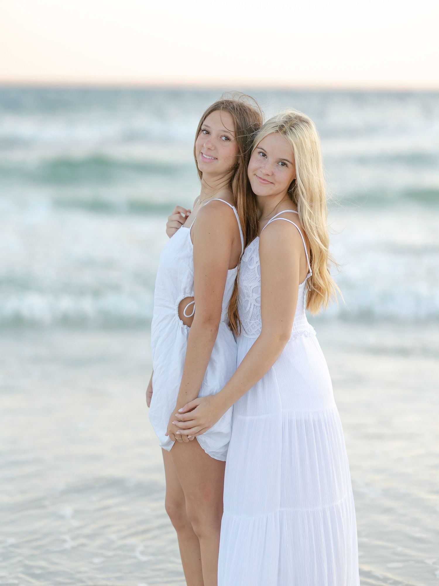 Friends in white dresses pose back-to-back on sunny beach.