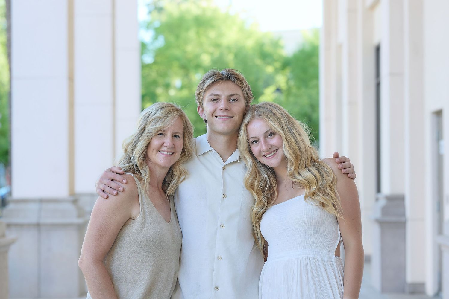 Three people in light-colored outfits smile together outdoors in front of a white column.