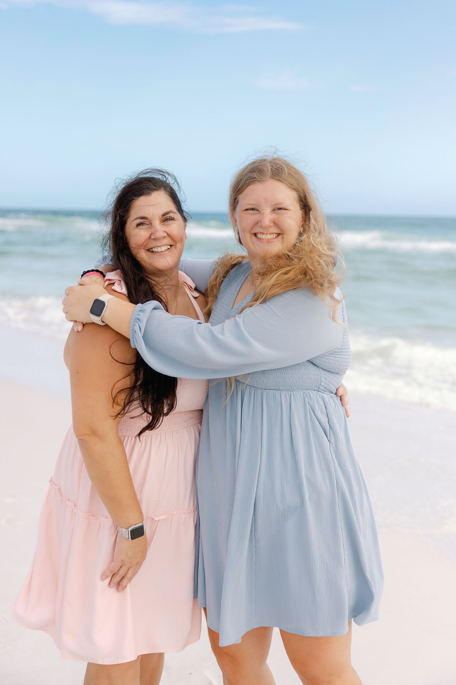 Two friends in flowy dresses laughing and embracing on a sunny beach with ocean waves in the background.