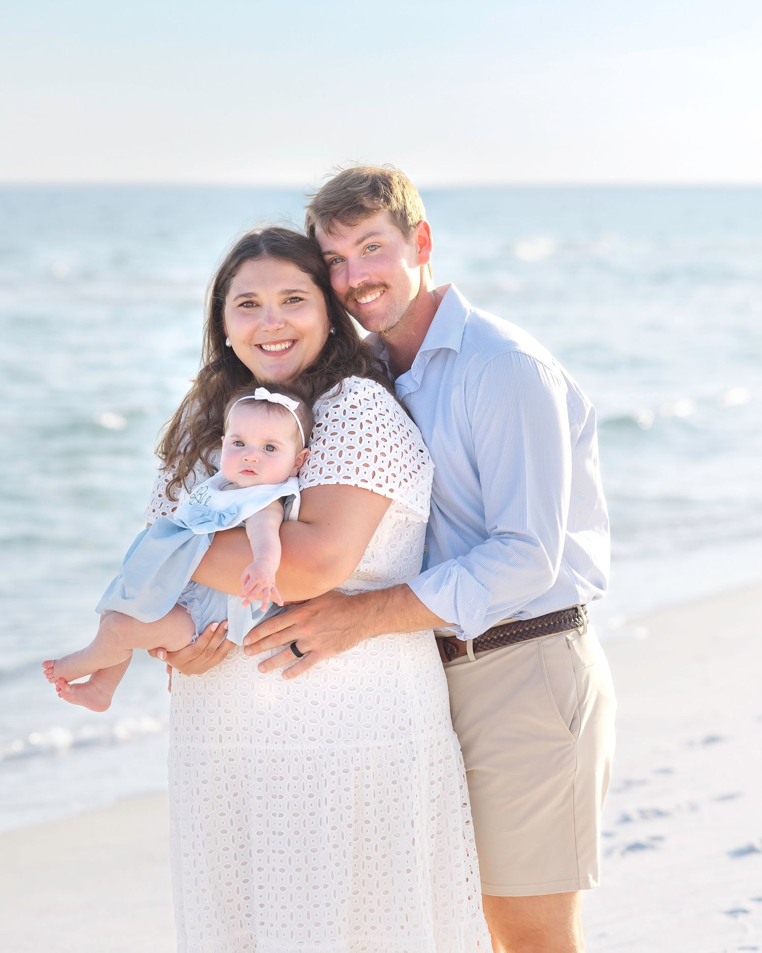 A family enjoys a tender moment together on a bright sandy beach with ocean waves in the background.