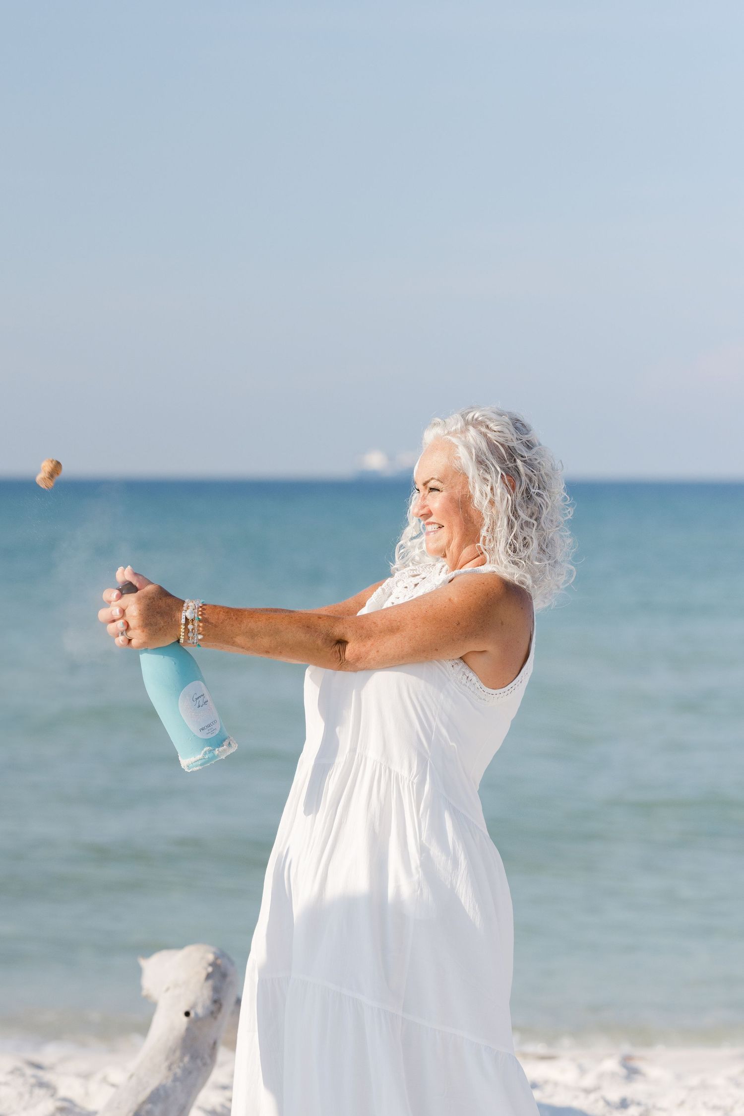 Person in flowing white dress joyfully holding water bottle on sunny beach with ocean view.