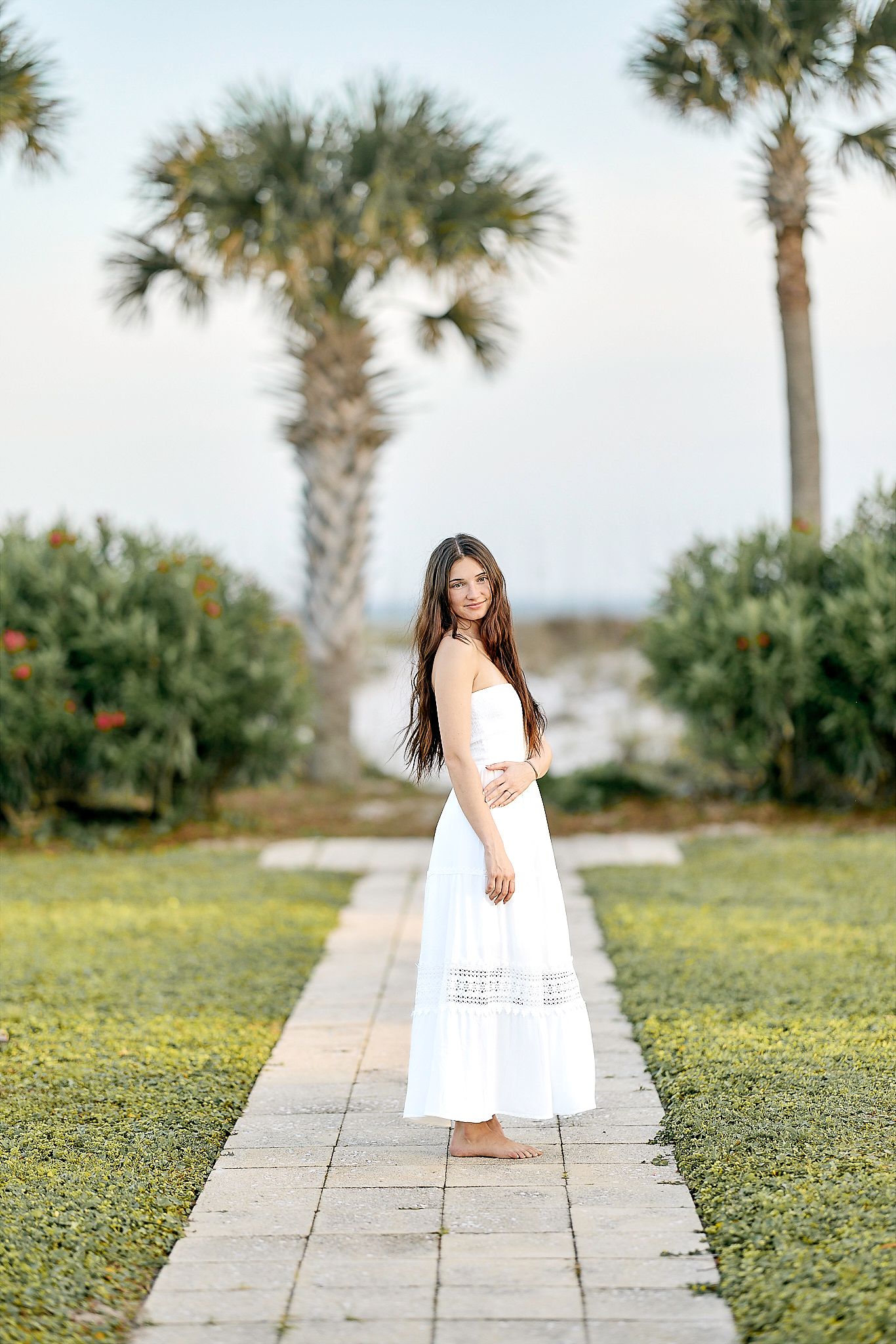 A white sundress flows elegantly on a stone path lined with palm trees and lush greenery in a tropical garden setting.