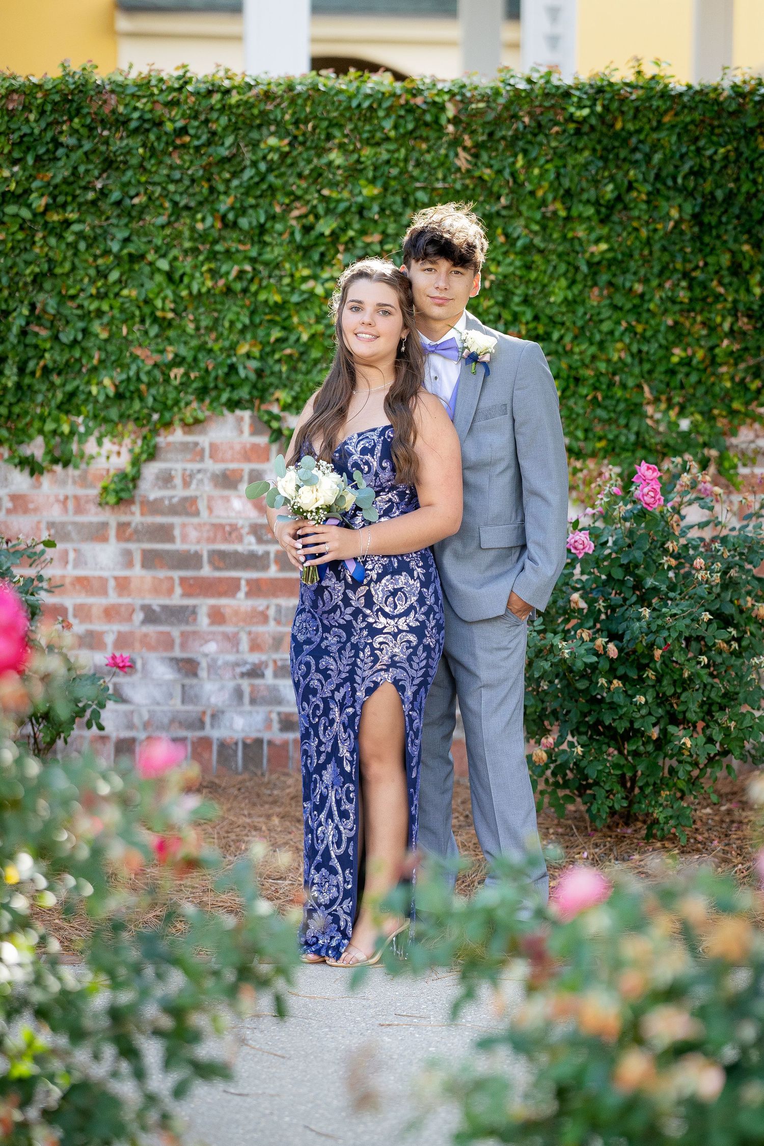 Couple in navy dress and gray suit pose together in garden with blooming rose bushes and brick wall background.