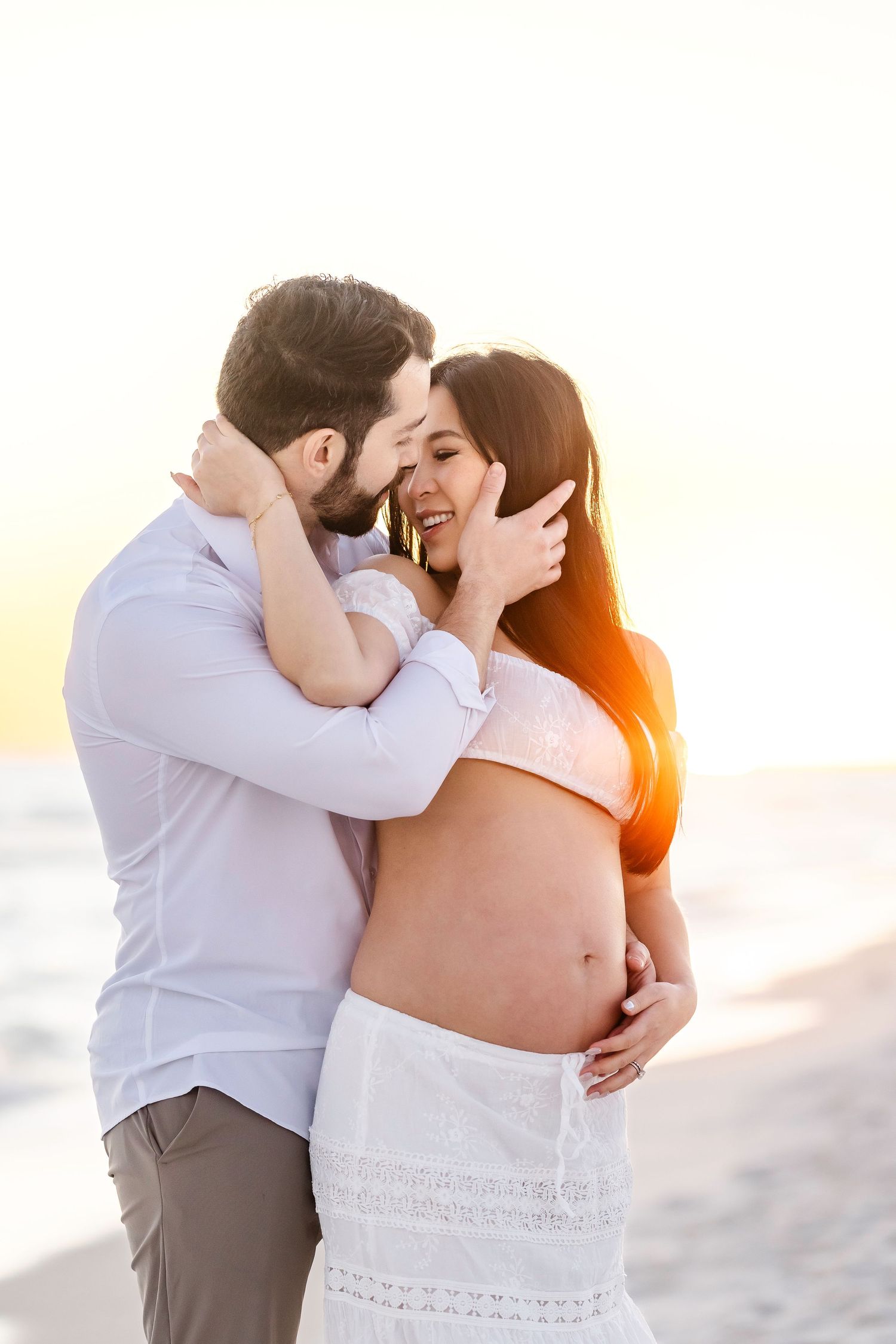 A couple embraces tenderly during a sunset beach photoshoot wearing white and beige attire.