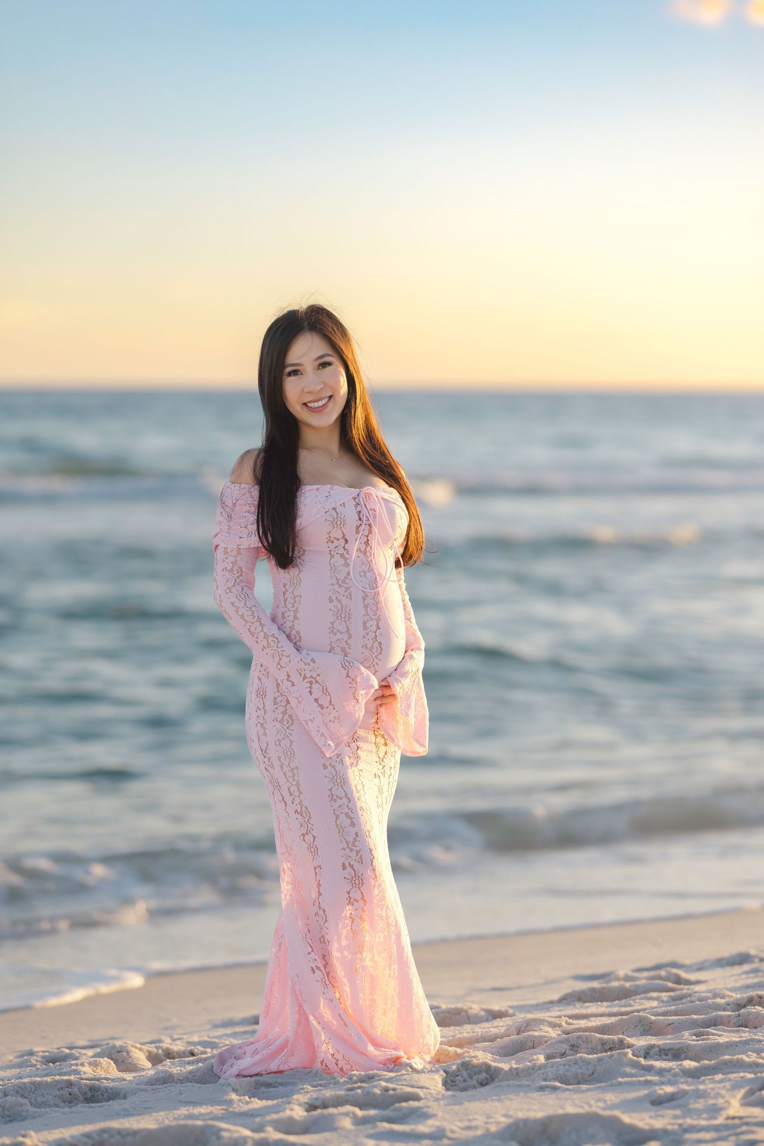 A woman in a pink lace dress poses on a pristine beach at sunset with ocean waves in the background.