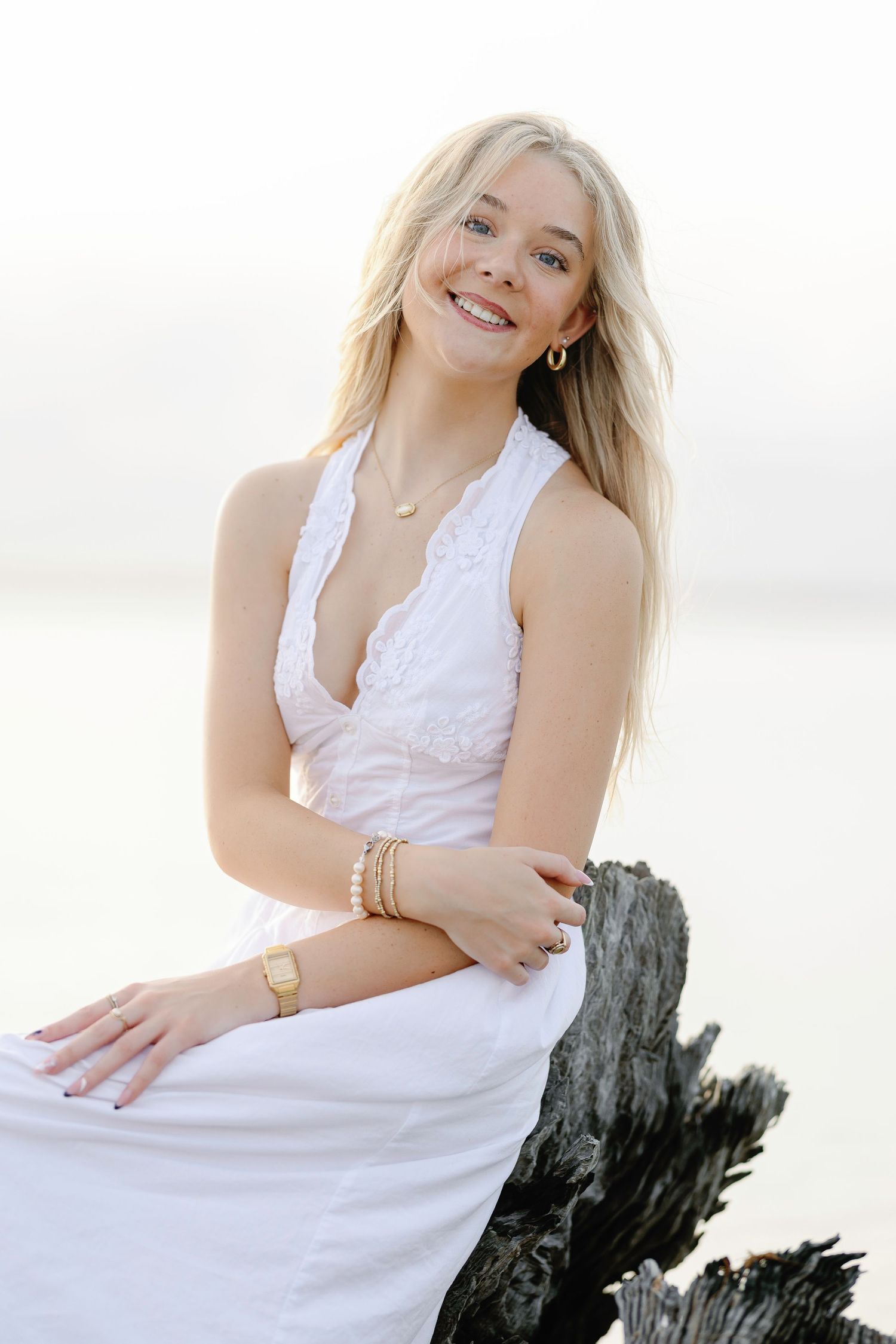 Person in white lace dress posing against weathered driftwood on a bright beach setting.