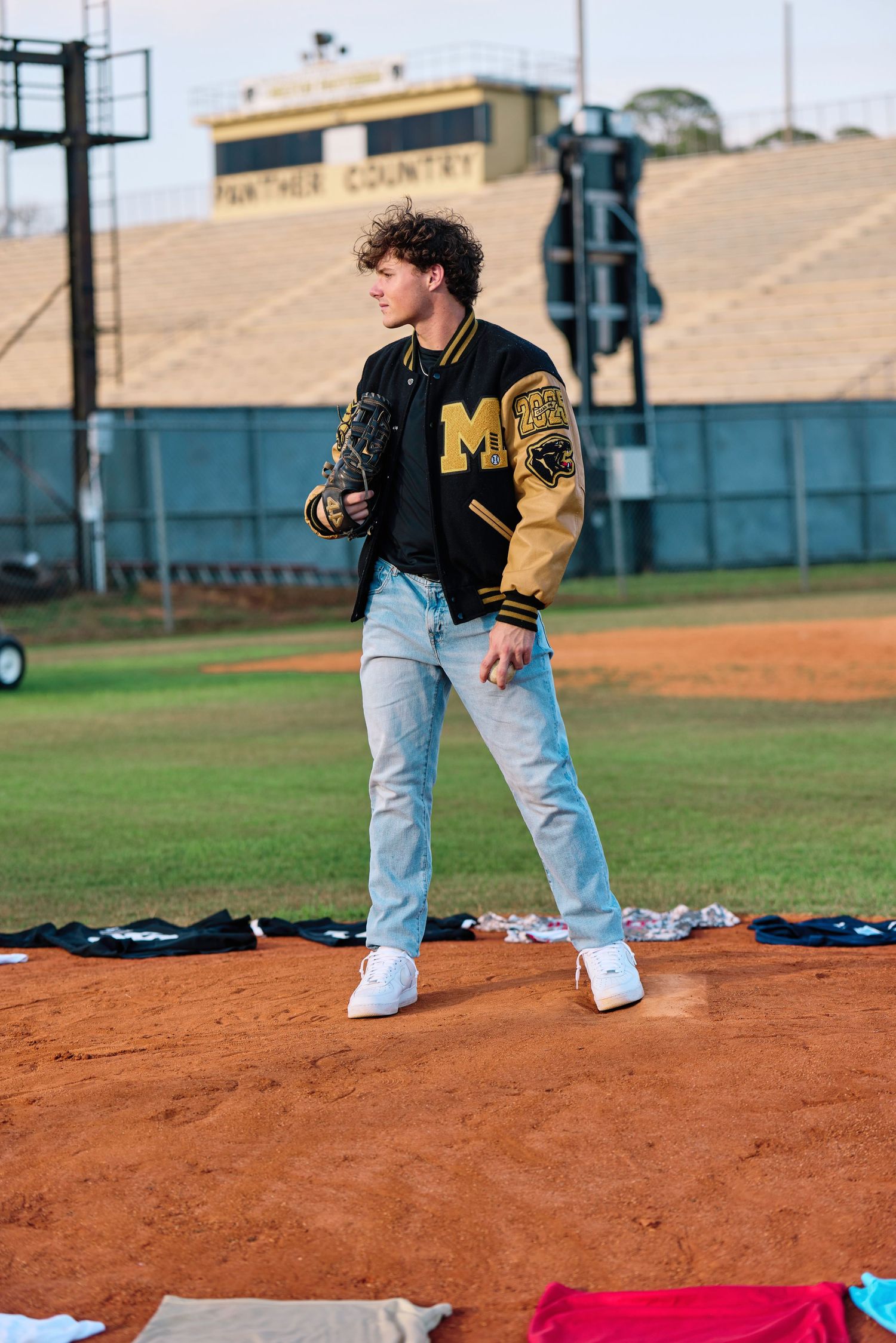 A baseball player in black and yellow varsity jacket stands on a dirt infield during practice.