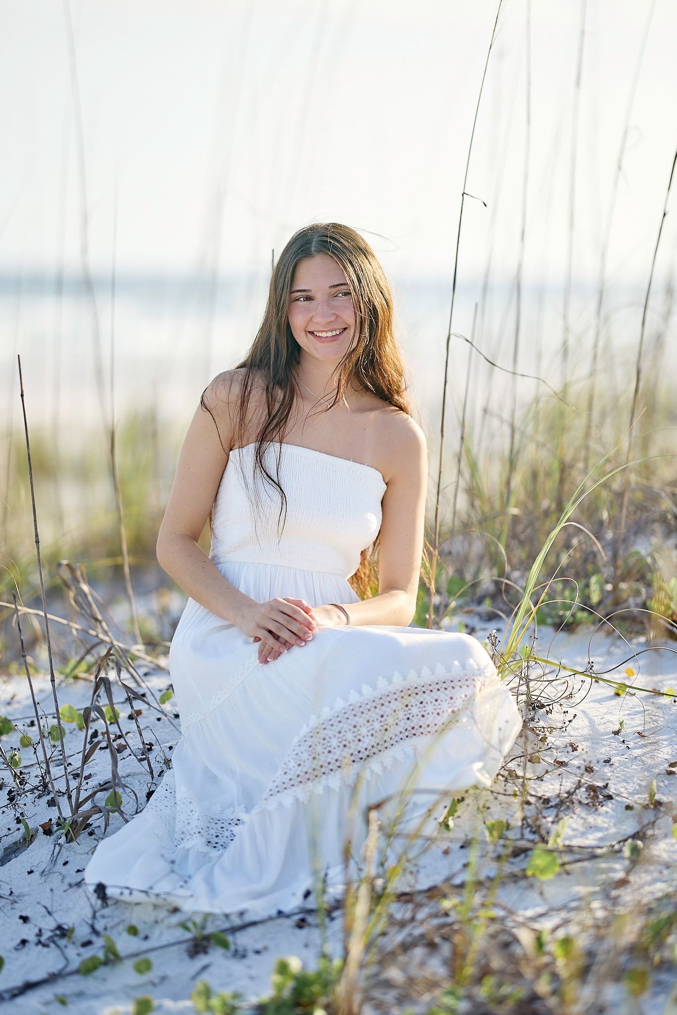 A white sundress glows in the soft light as someone poses among beach grass and sand dunes near the ocean.