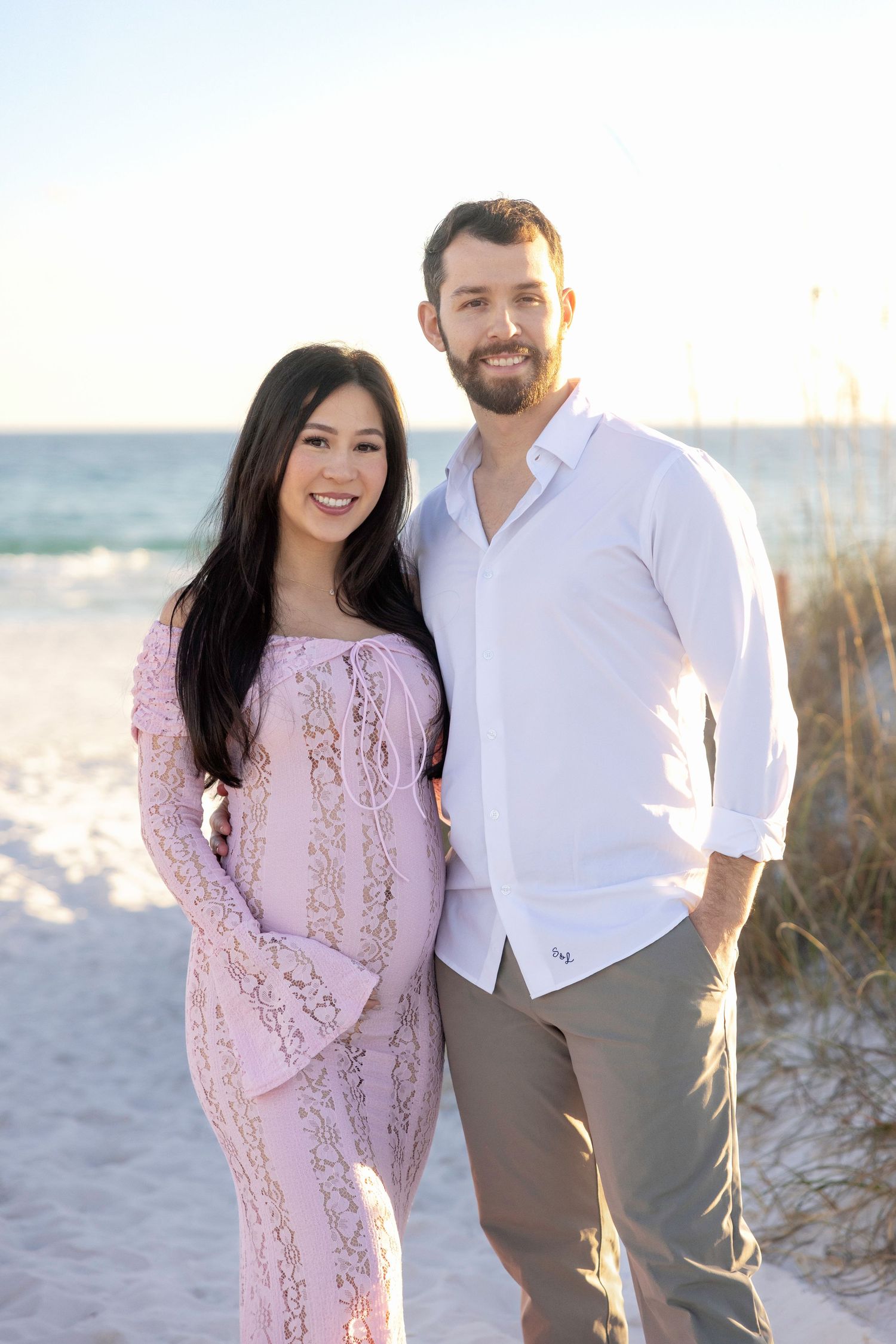 A couple in a pink lace dress and white shirt pose together on a sunny beach with sand dunes in the background.