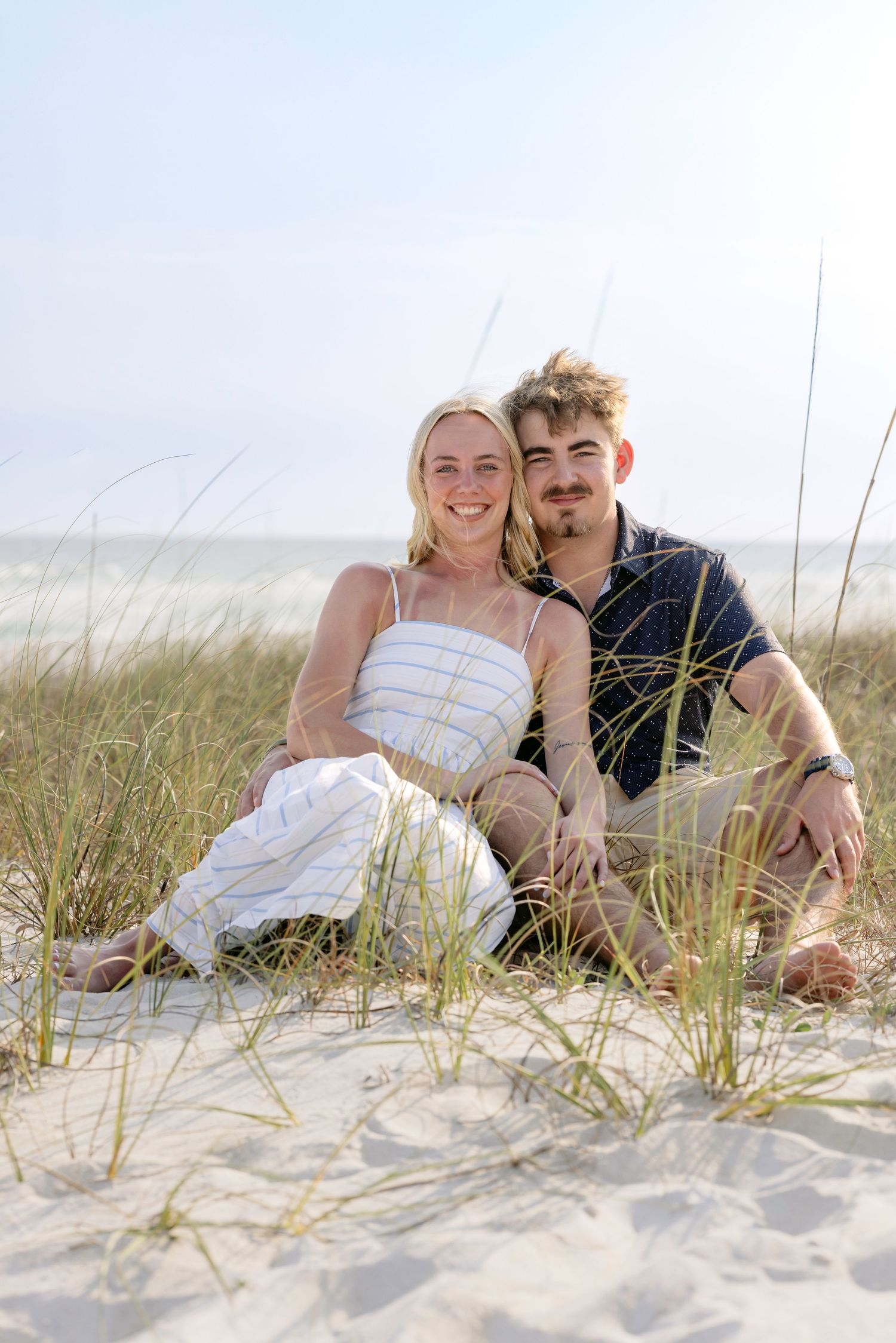 A young couple sitting together on sandy beach dunes with sea grass, smiling at the camera on a sunny day.