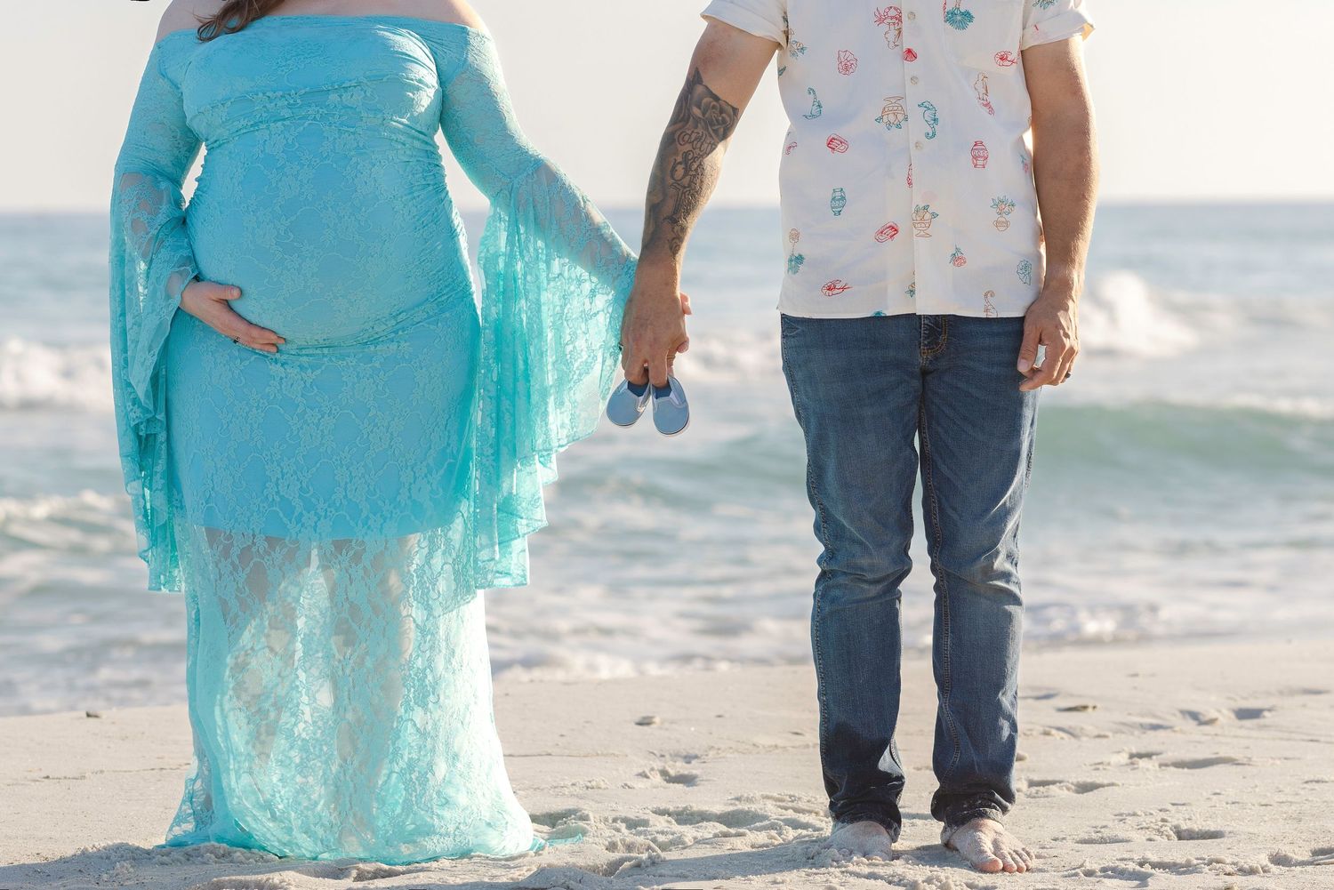 Pregnant couple holding baby shoes on beach, woman in turquoise lace maternity gown, man in jeans and floral shirt.