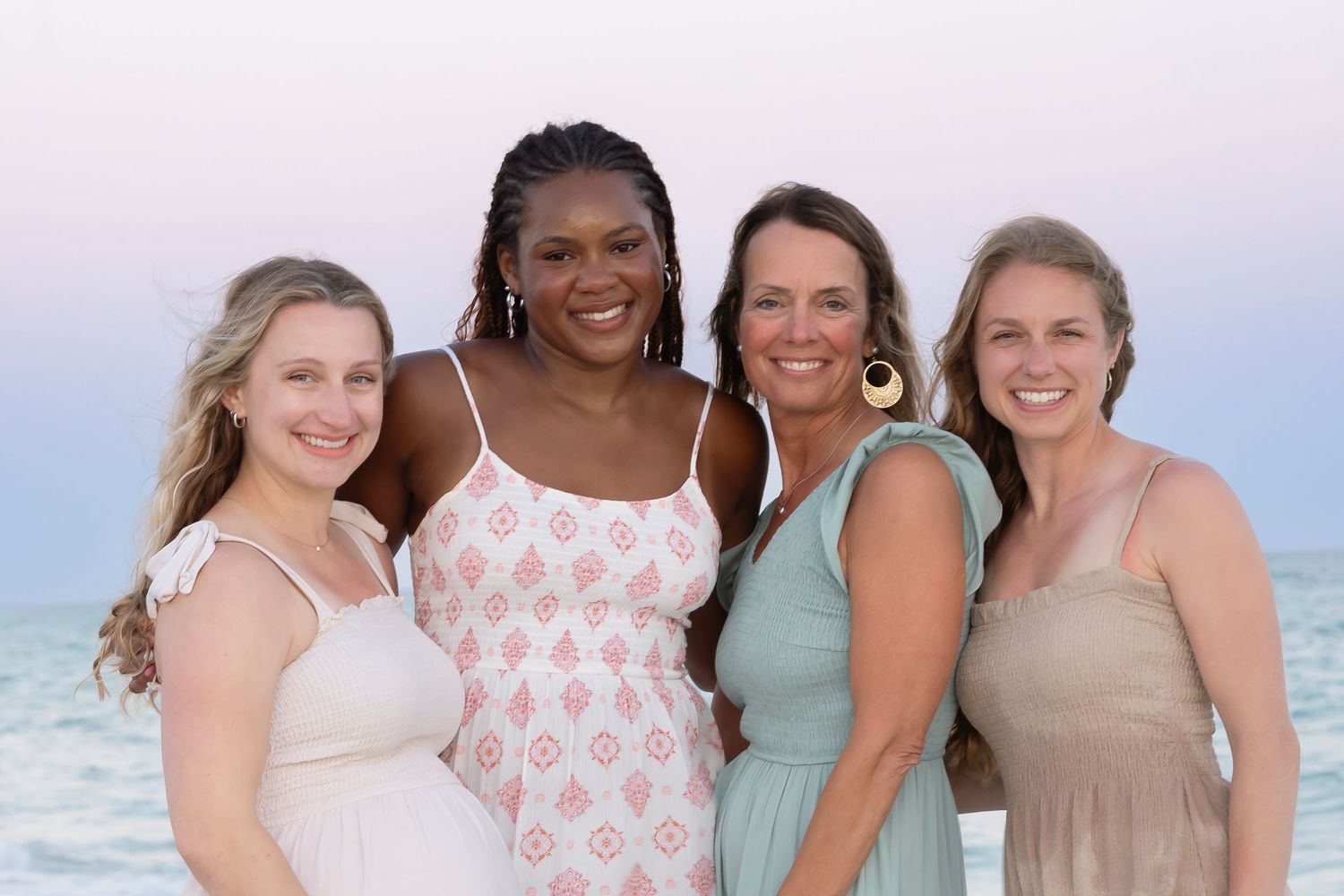Four smiling women posing together on a beach at dusk, dressed in elegant pastel outfits with ocean backdrop.