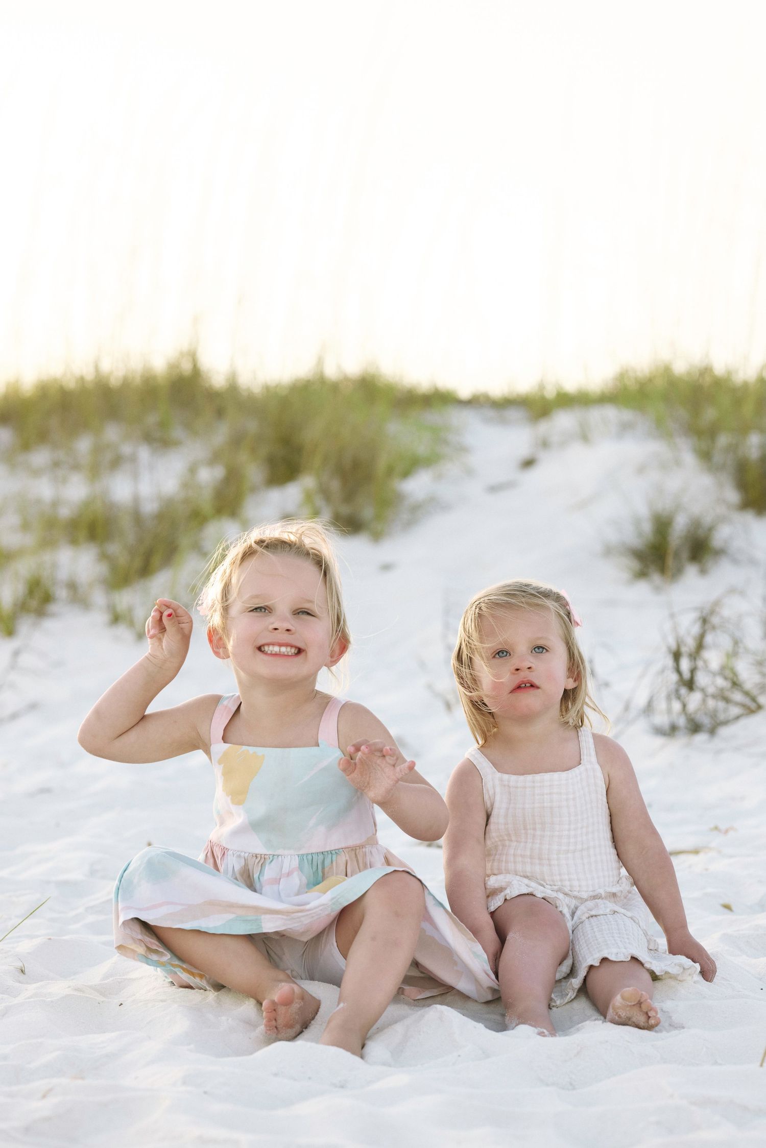 Two young blonde sisters in summer dresses sit together on white sand beach near sea oat dunes.