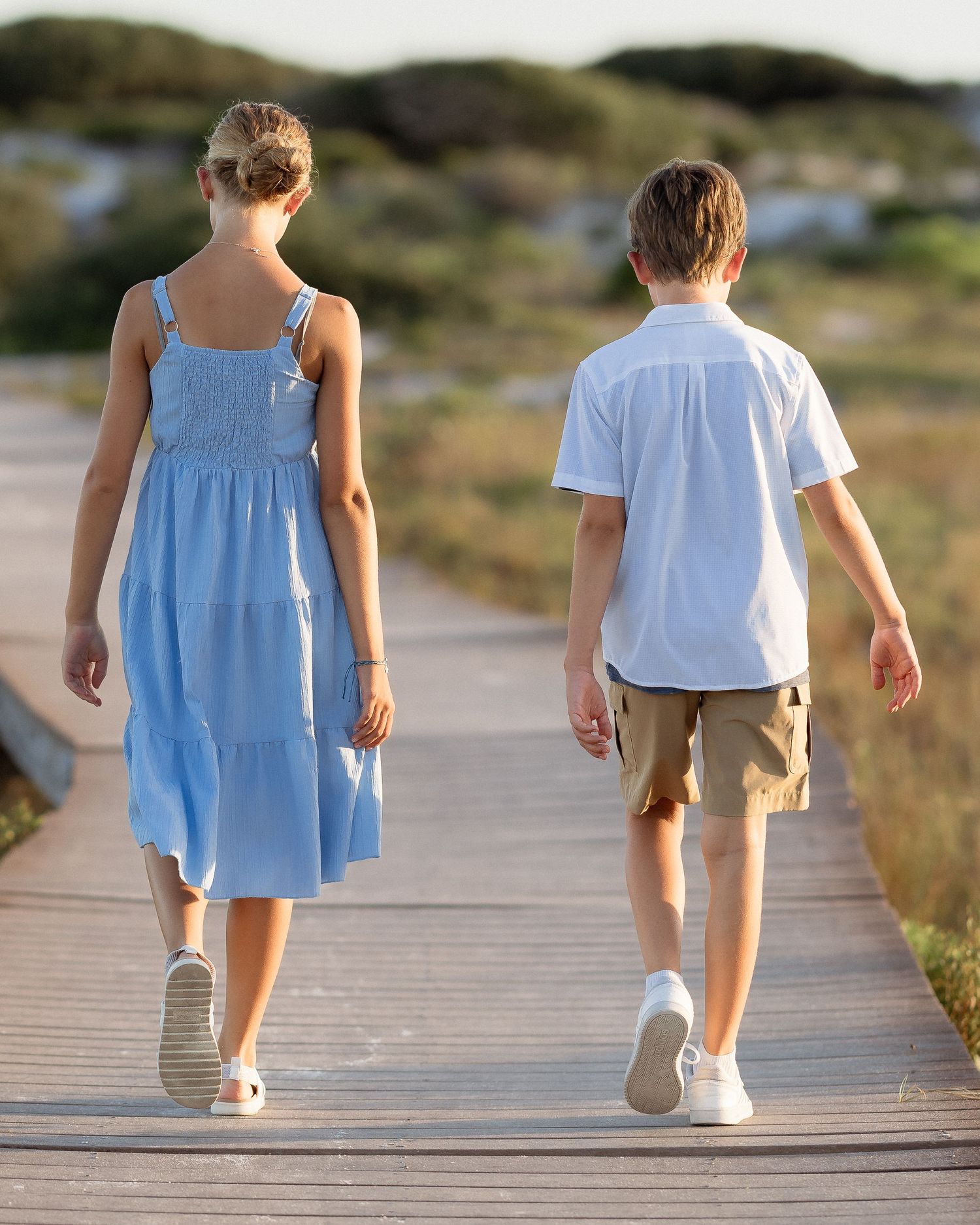 Two children walk hand in hand on a wooden boardwalk, girl in blue dress and boy in white shirt and khaki shorts.