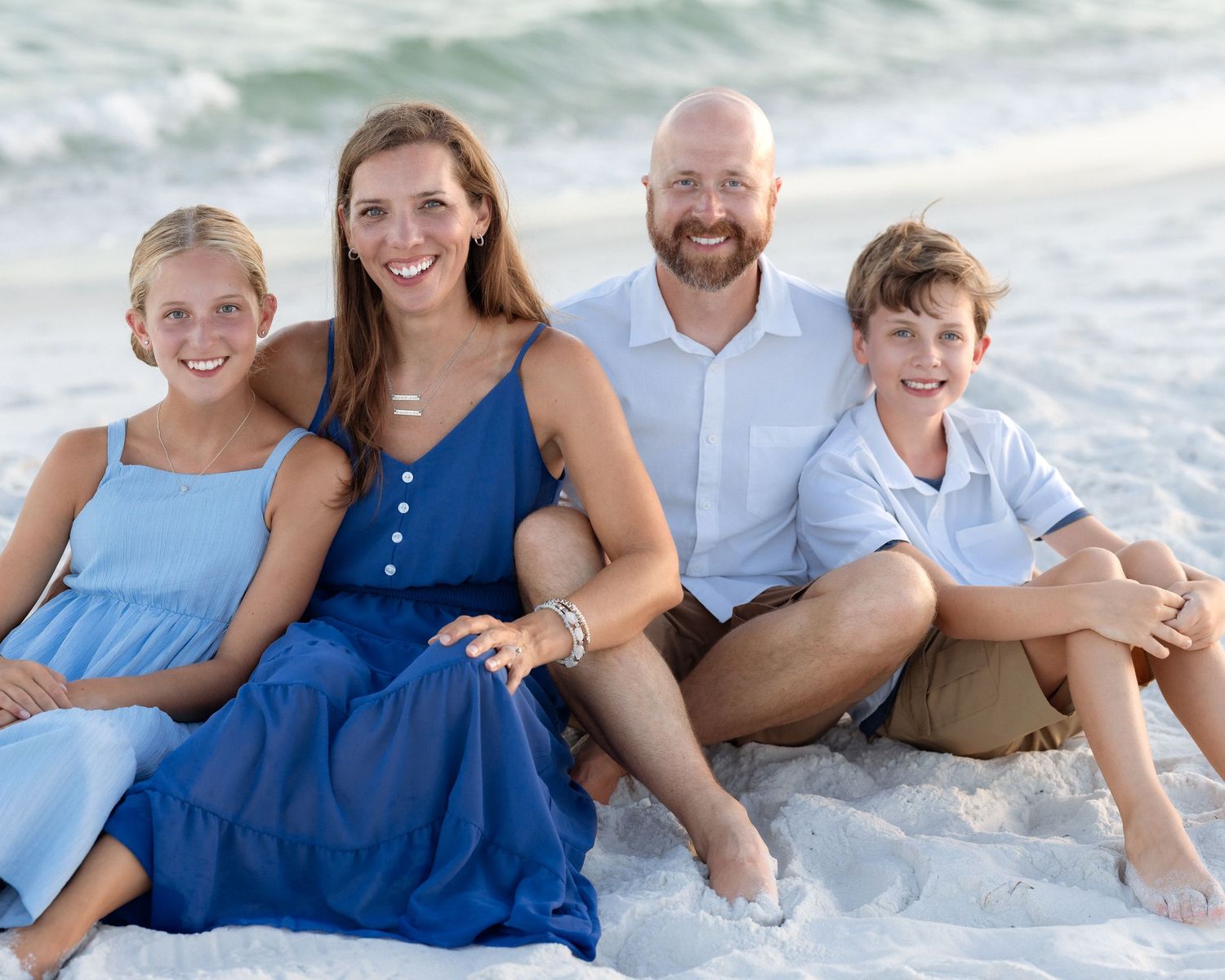 Family of four sitting together on sandy beach shoreline, smiling in coordinated blue and white outfits.