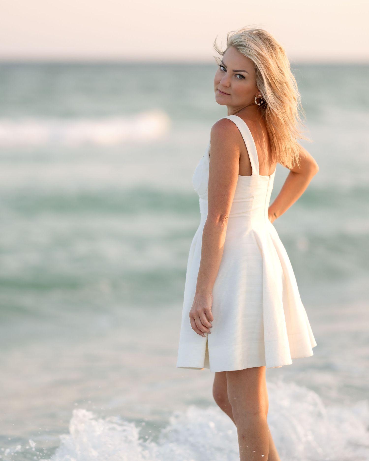 Blonde woman in white fit-and-flare dress poses at ocean's edge during golden hour.