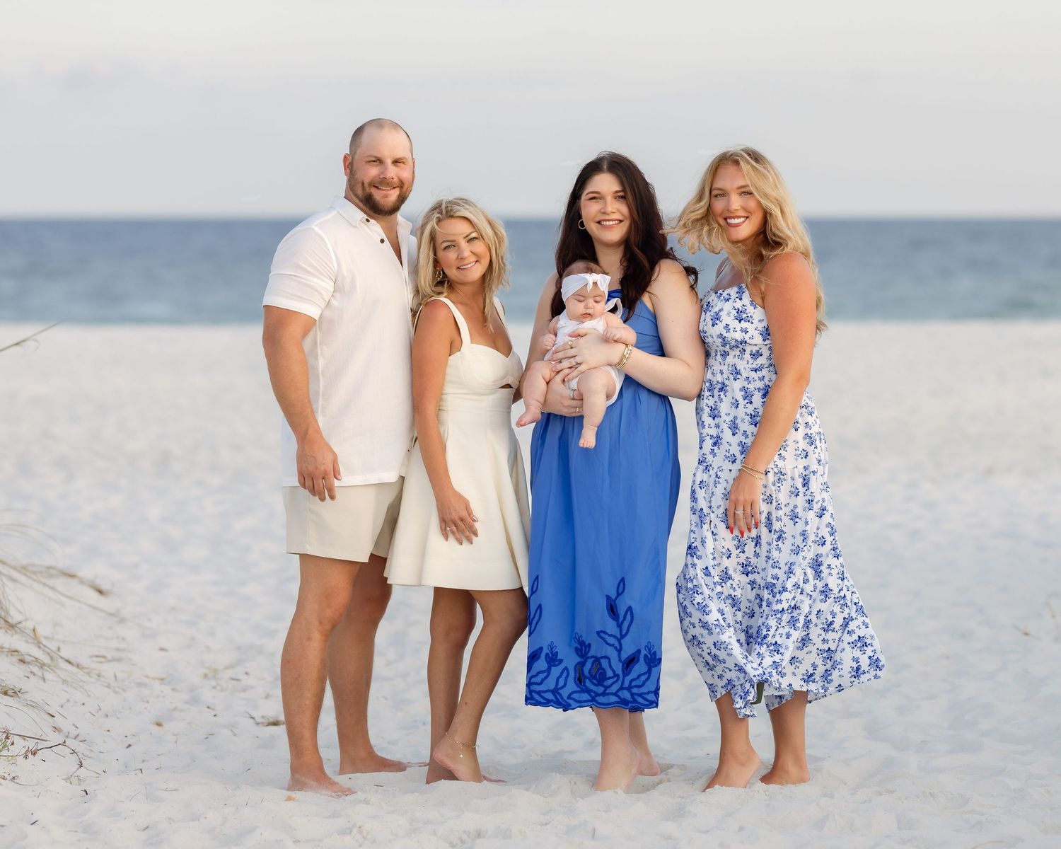 Family of four posing on a white sand beach, with a woman in blue holding a baby, on a sunny day.