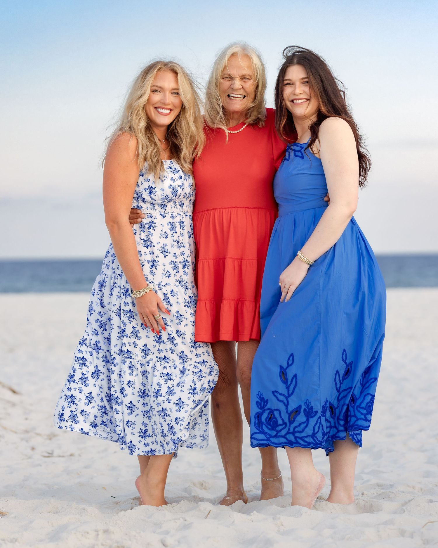 Three women in colorful dresses smile together on a sunny beach, wearing white, red, and blue outfits.