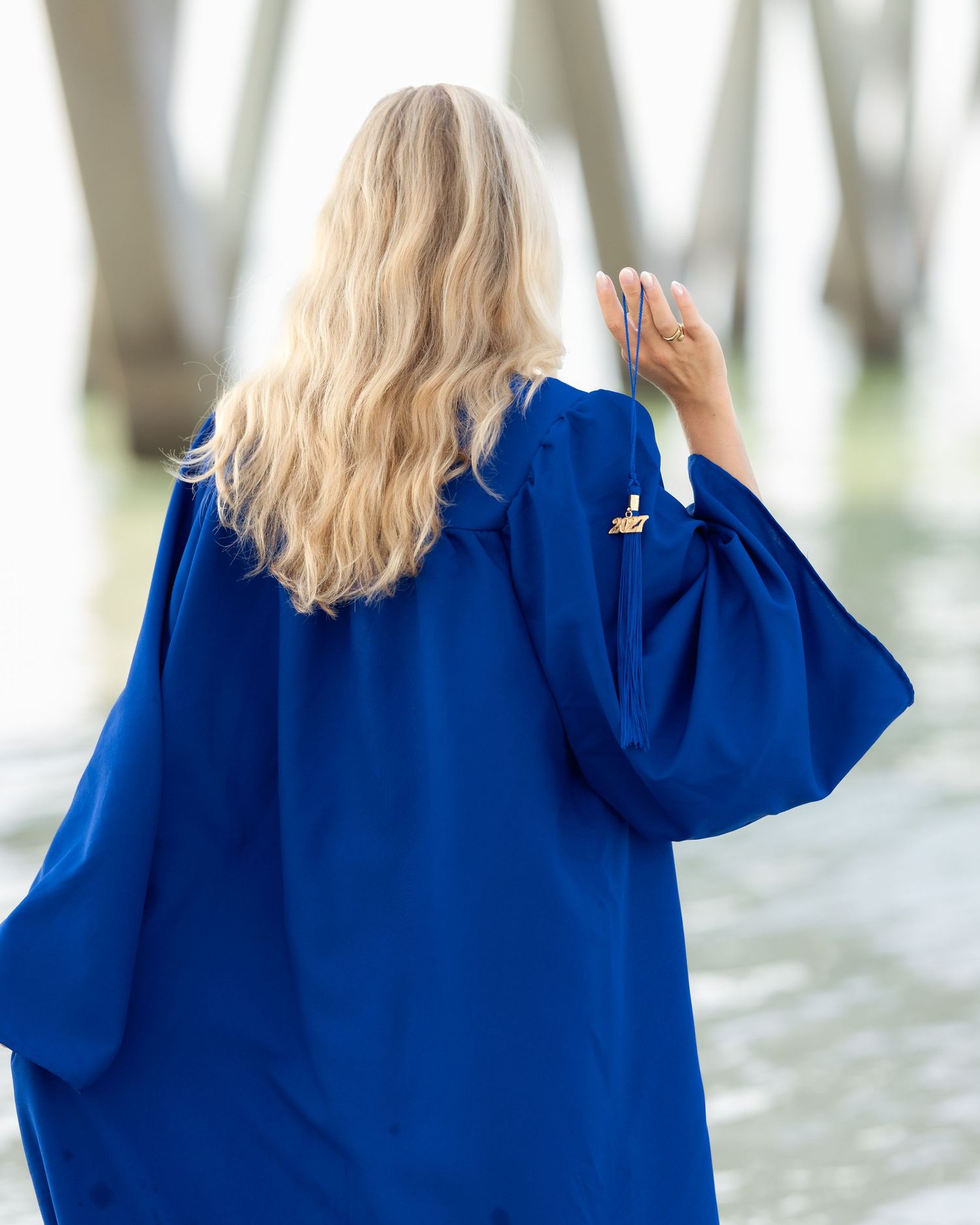 Blonde woman in royal blue graduation gown standing near a pier by the water, viewed from behind.