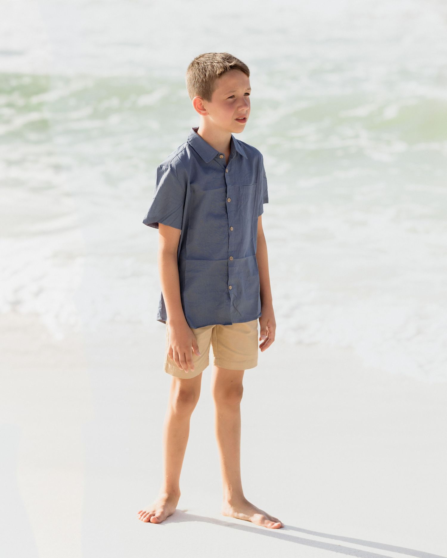 Young boy in blue shirt and khaki shorts stands barefoot on sandy beach with waves.