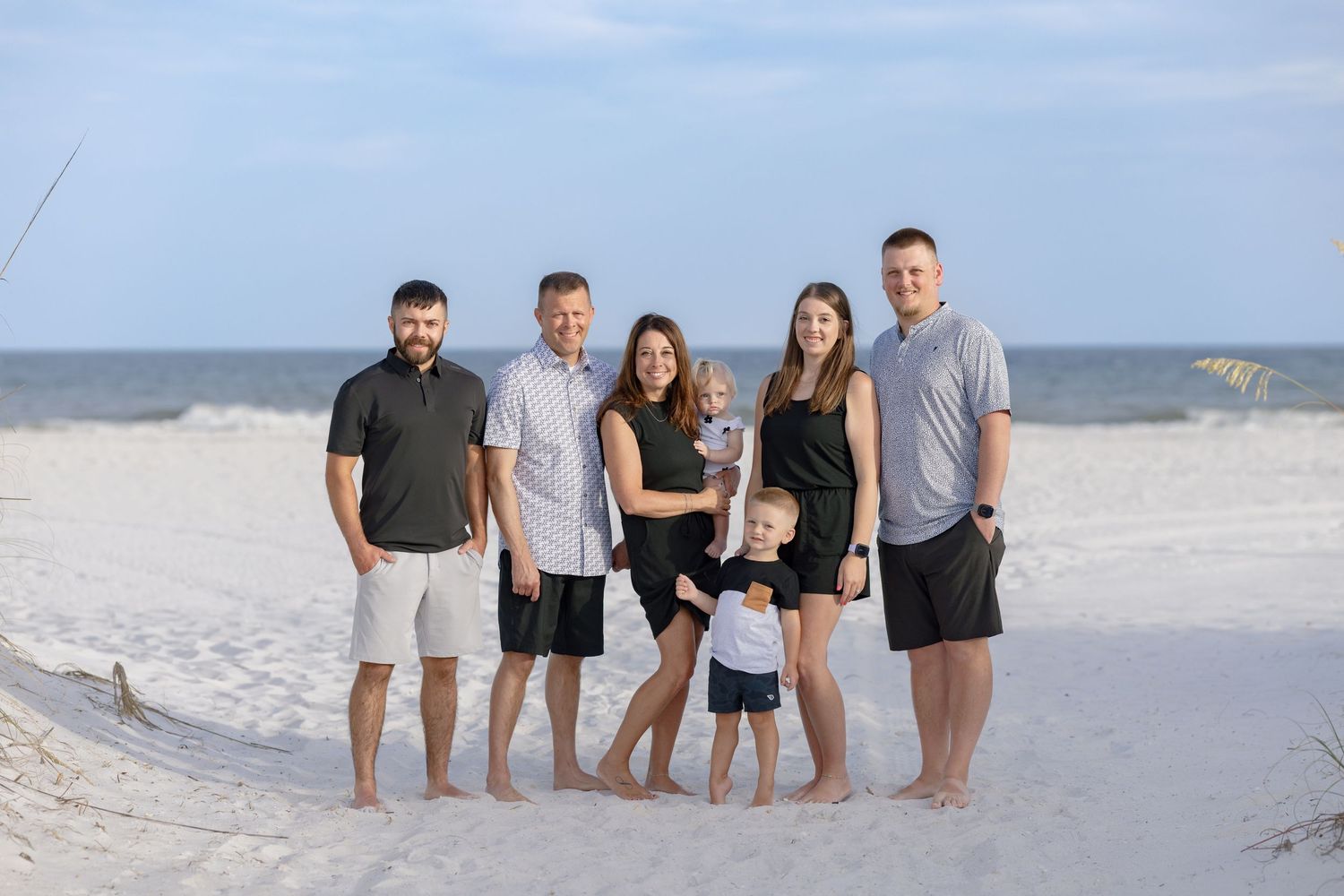 Family of six posing together on a white sandy beach with calm ocean waves in the background on a clear day.