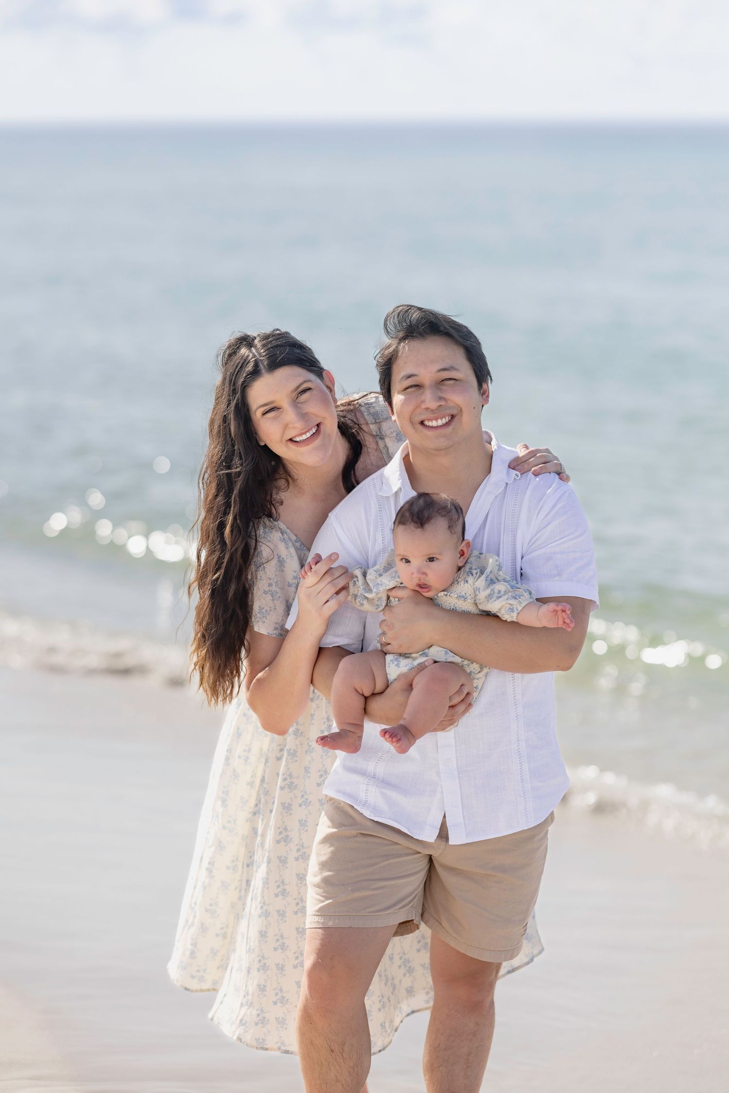 Happy young family with newborn baby posing on a sunny beach, with ocean waves in the background.