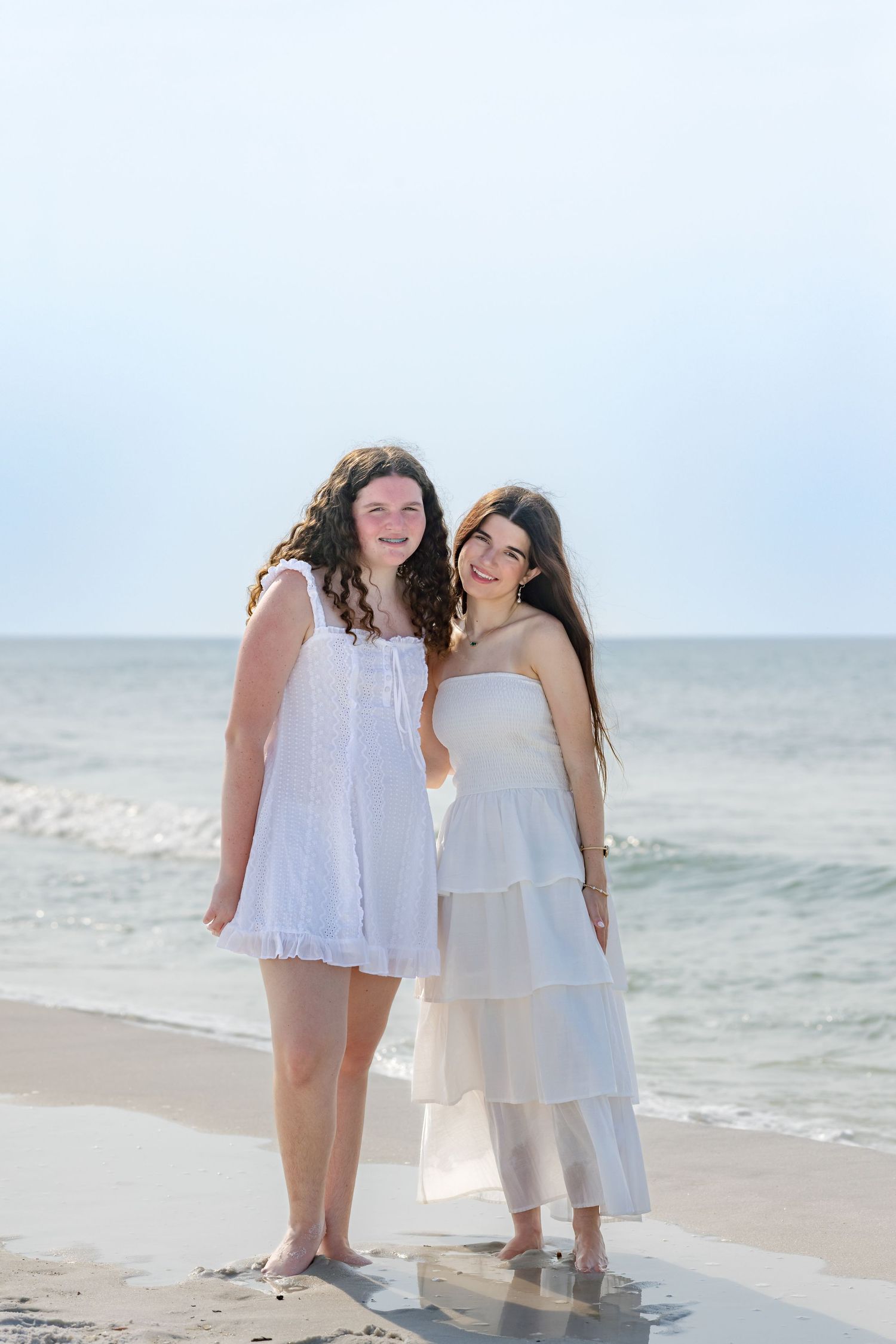 Two young women in white dresses smiling together on sunny beach with ocean behind them.