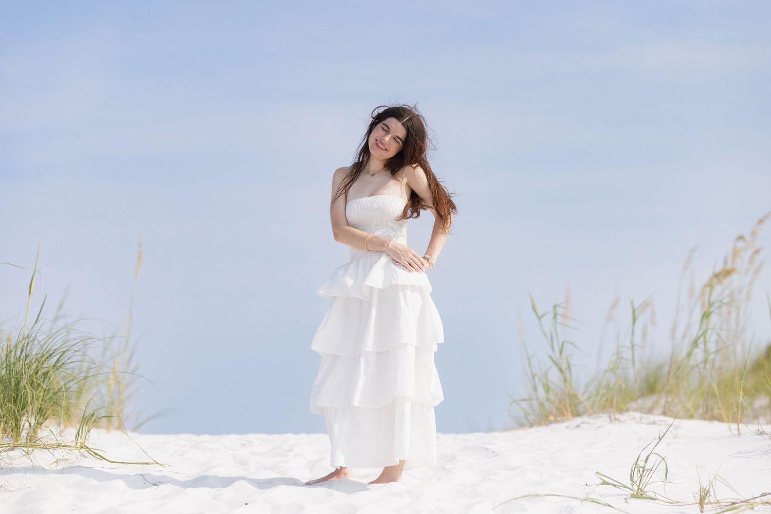 Woman in layered white tiered dress posing on white sand dunes with sea grass under blue sky.